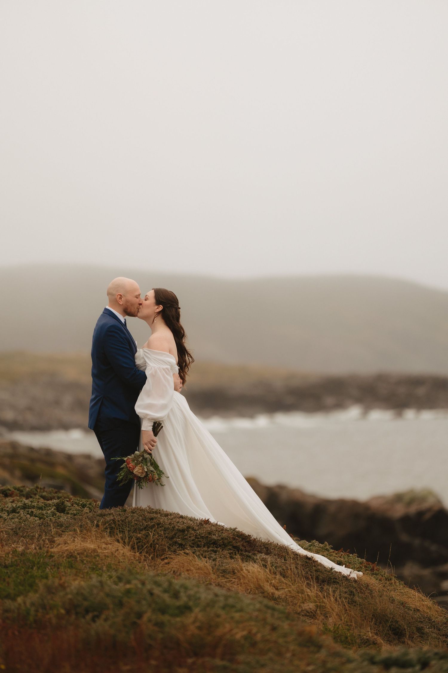 A couple shares a romantic moment on a misty coastal cliff in wedding attire against a moody landscape.