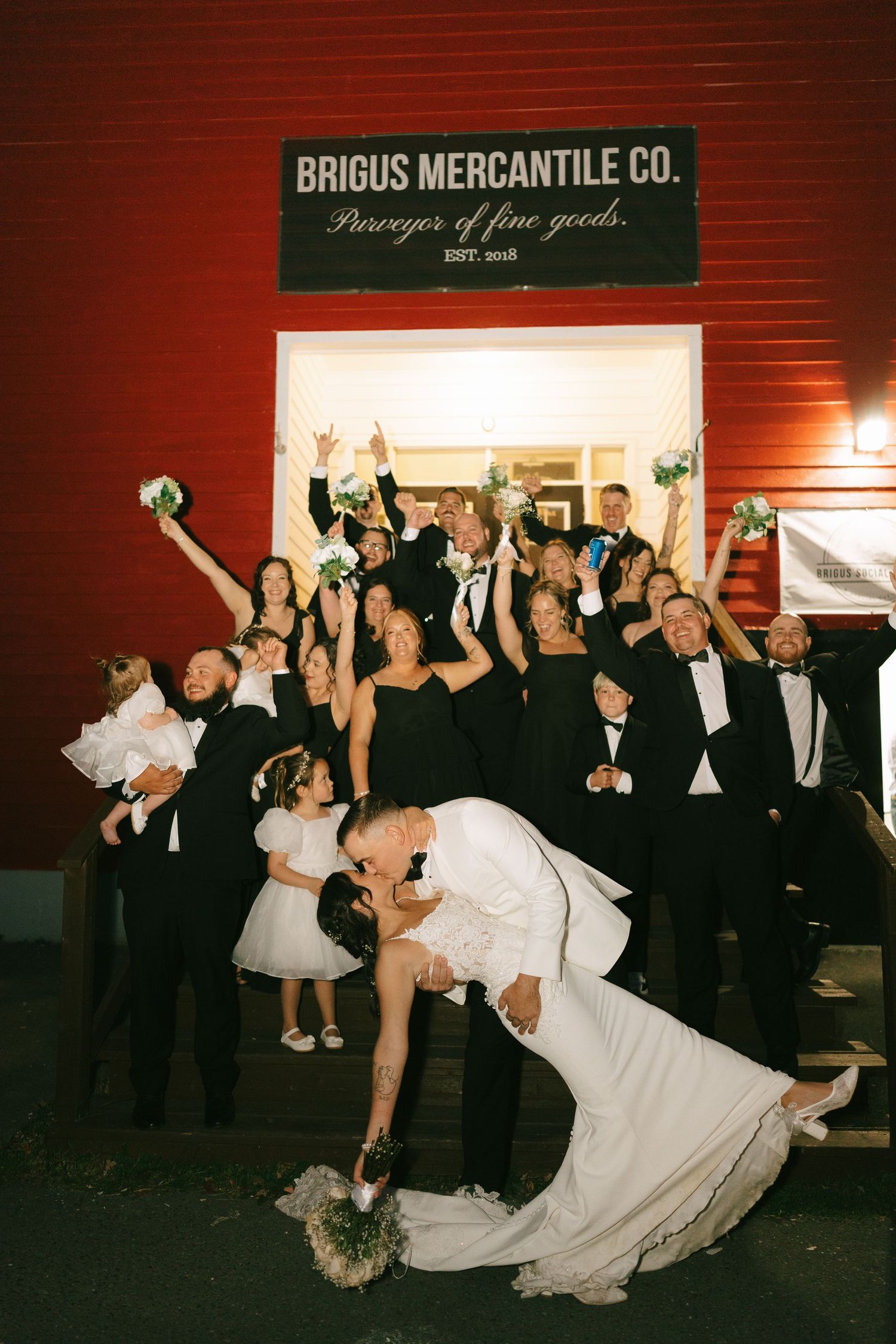 Wedding party celebrates with a dramatic dip pose in front of Brigus Mercantile Co. building at night.