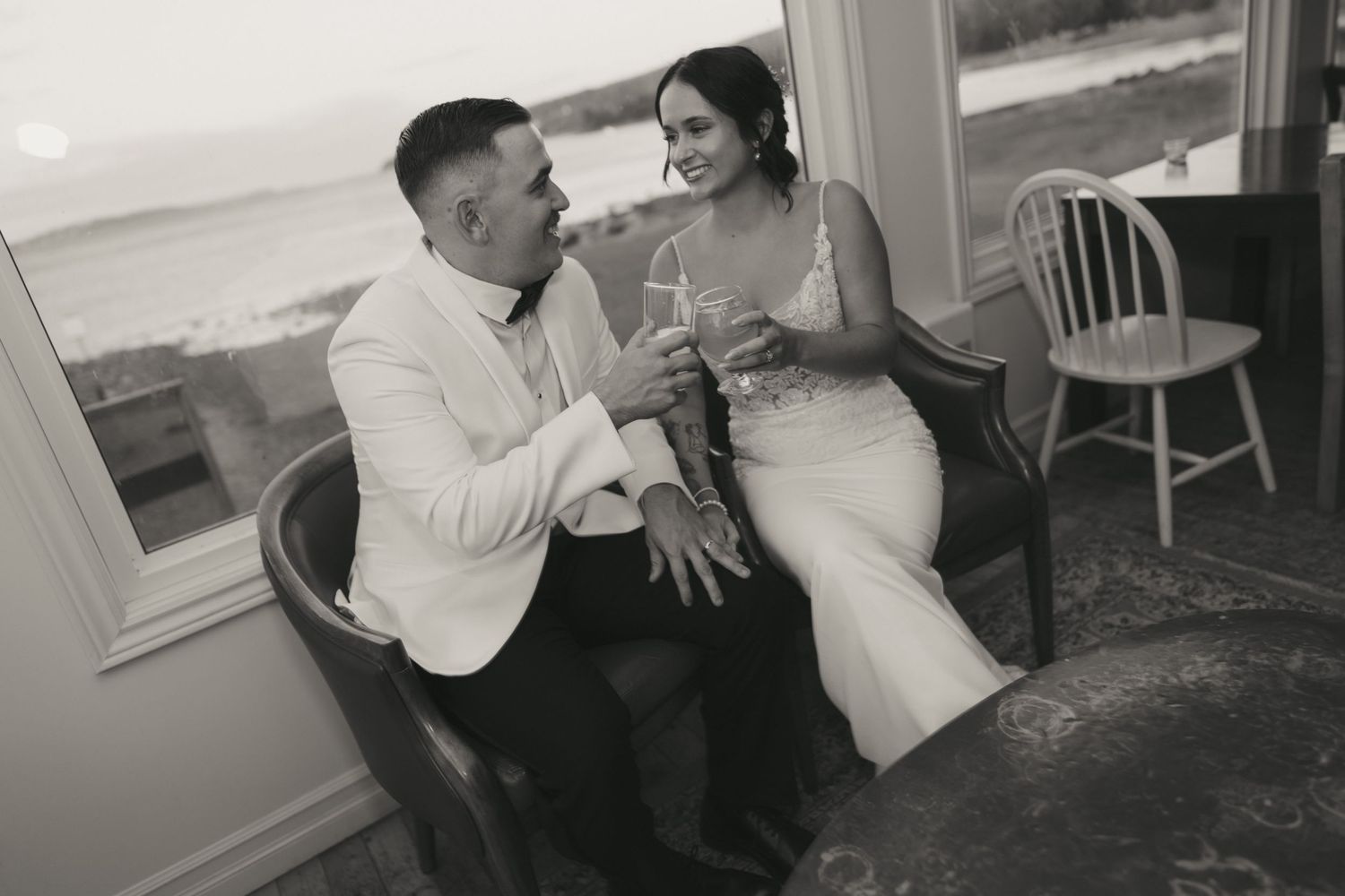 A couple shares an intimate moment while toasting with wine glasses at a seaside venue in black and white.