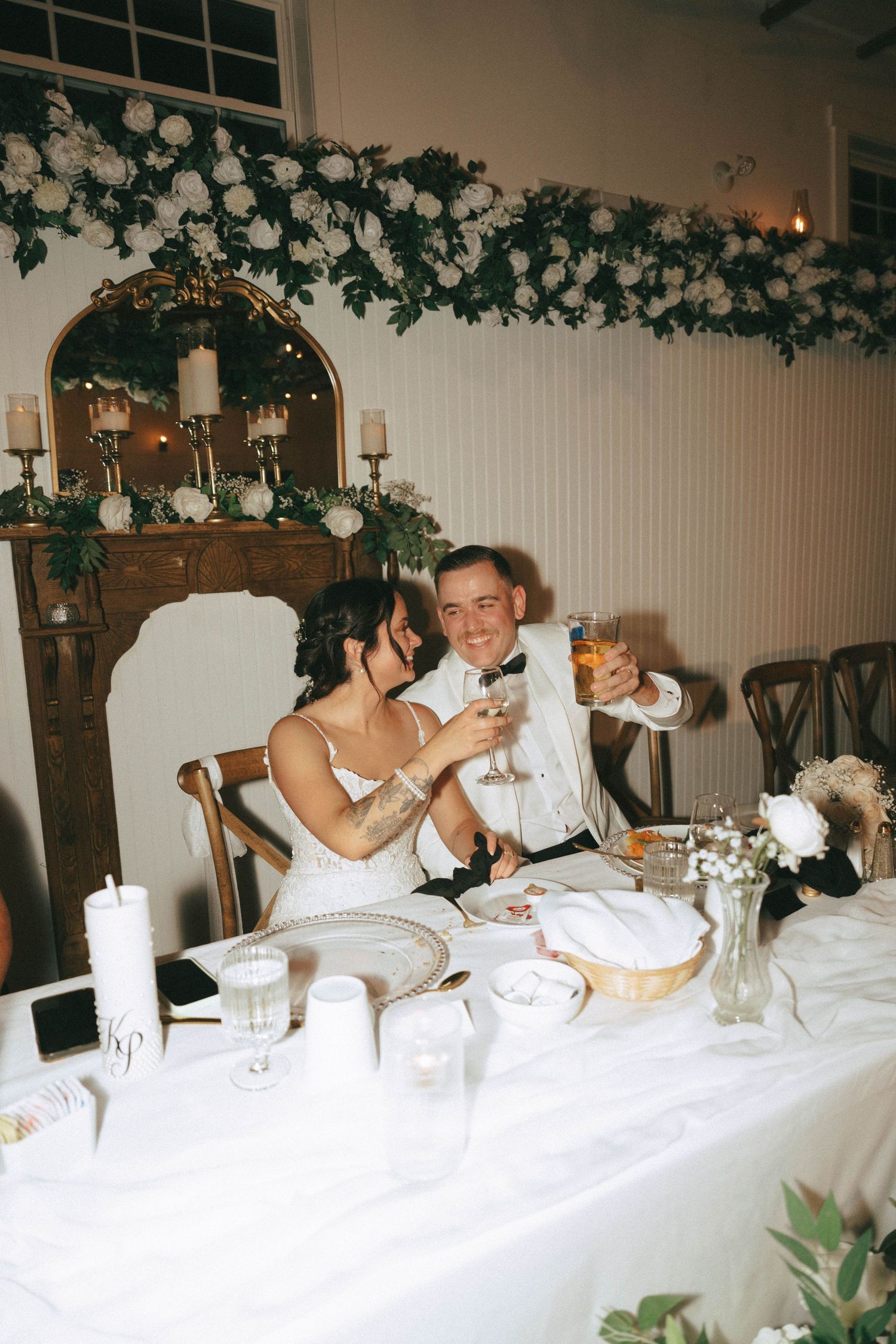 A couple toasts with champagne glasses at their wedding reception table decorated with white flowers and greenery.
