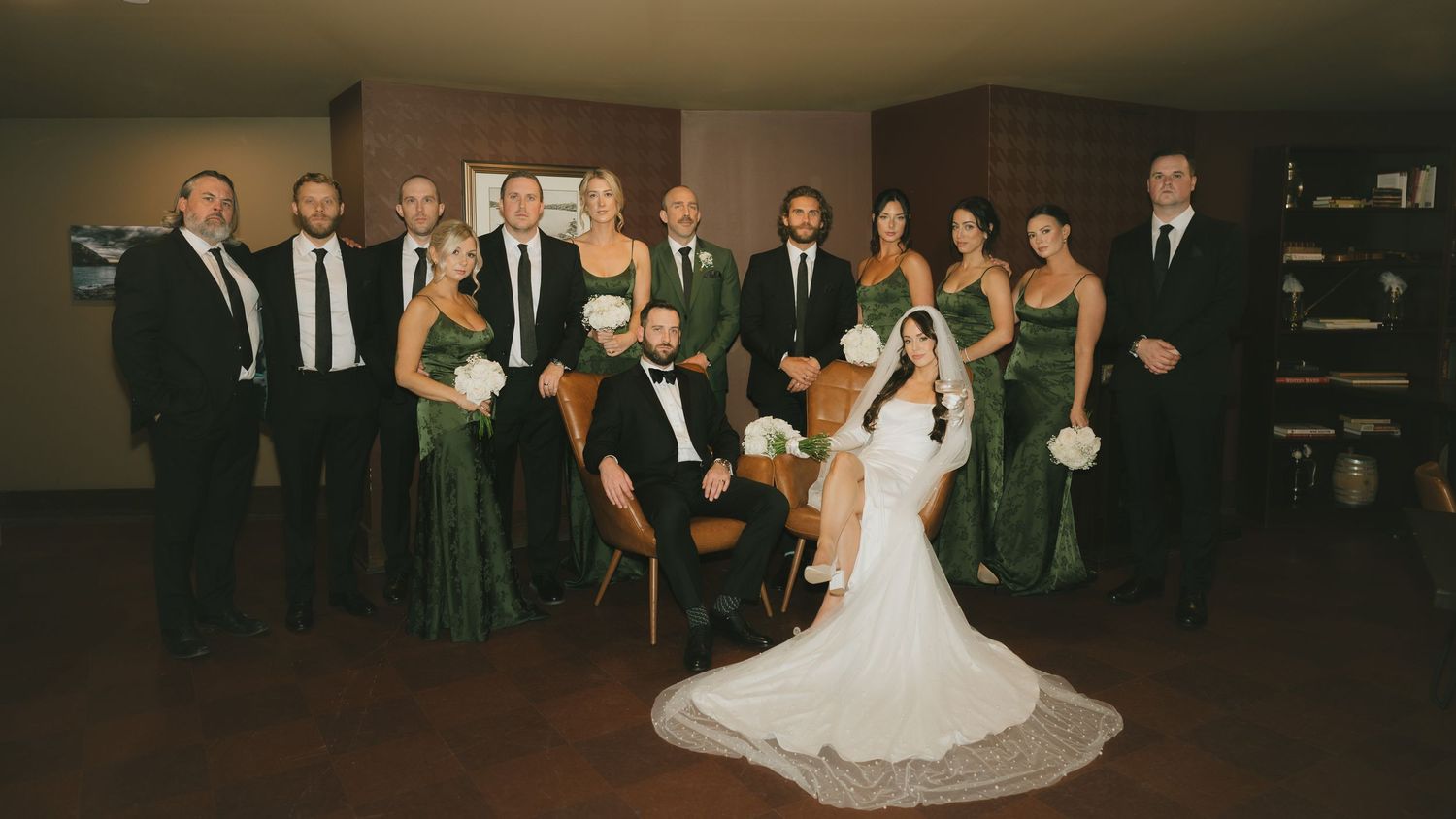 Wedding party in green and black attire poses with bride and groom in elegant indoor venue.