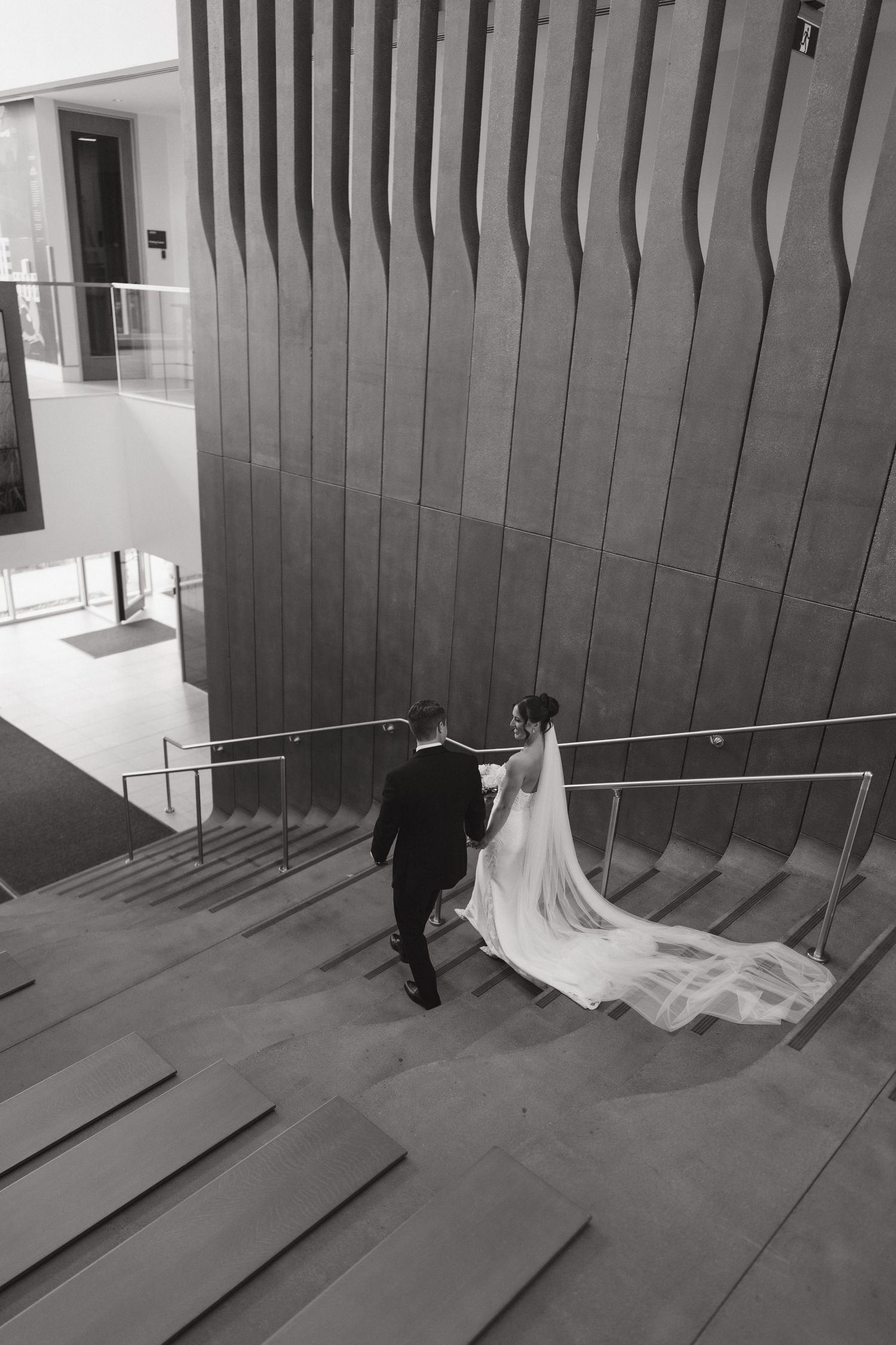 Black and white photo of bride and groom walking up stairs, bride's veil trailing behind, modern architectural venue.