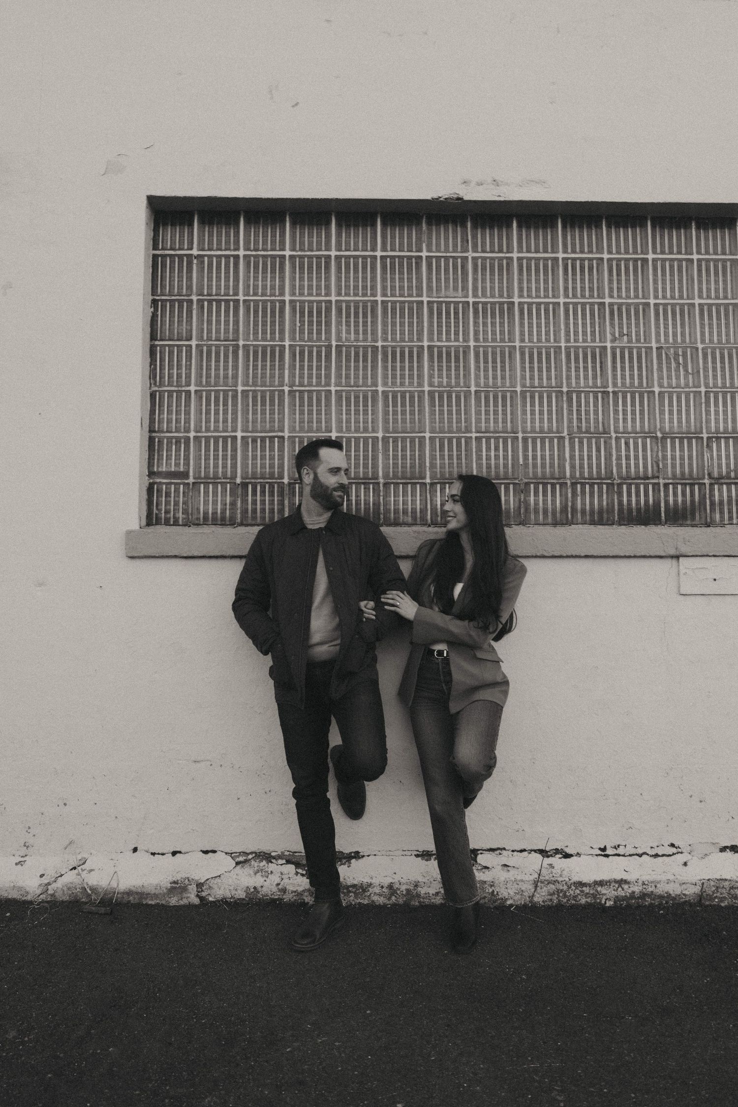 Black and white photo of a couple leaning against a wall, smiling at each other in a candid romantic moment.