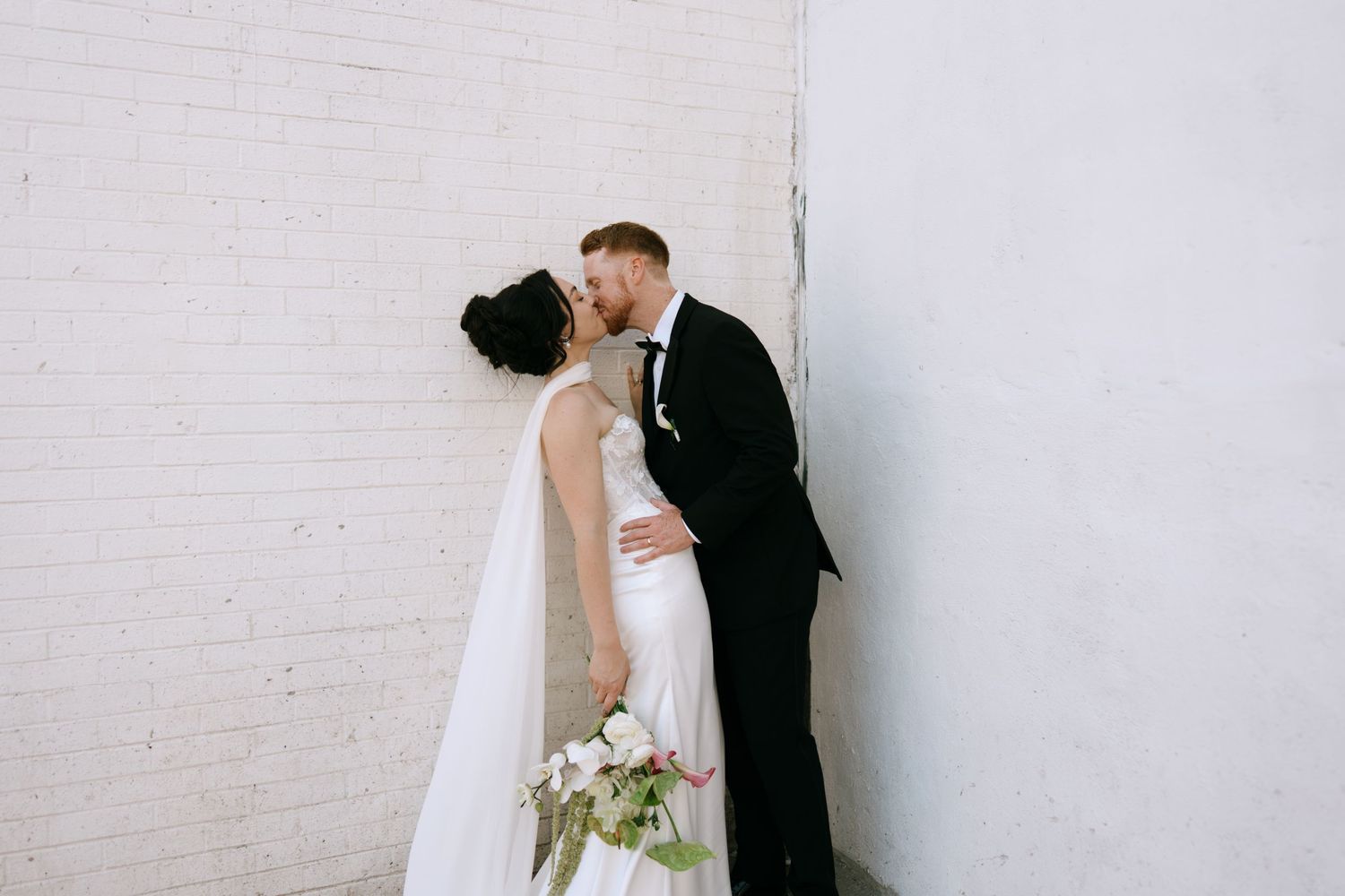 A bride and groom share a romantic kiss against a white brick wall, the bride holding a bouquet of white flowers.