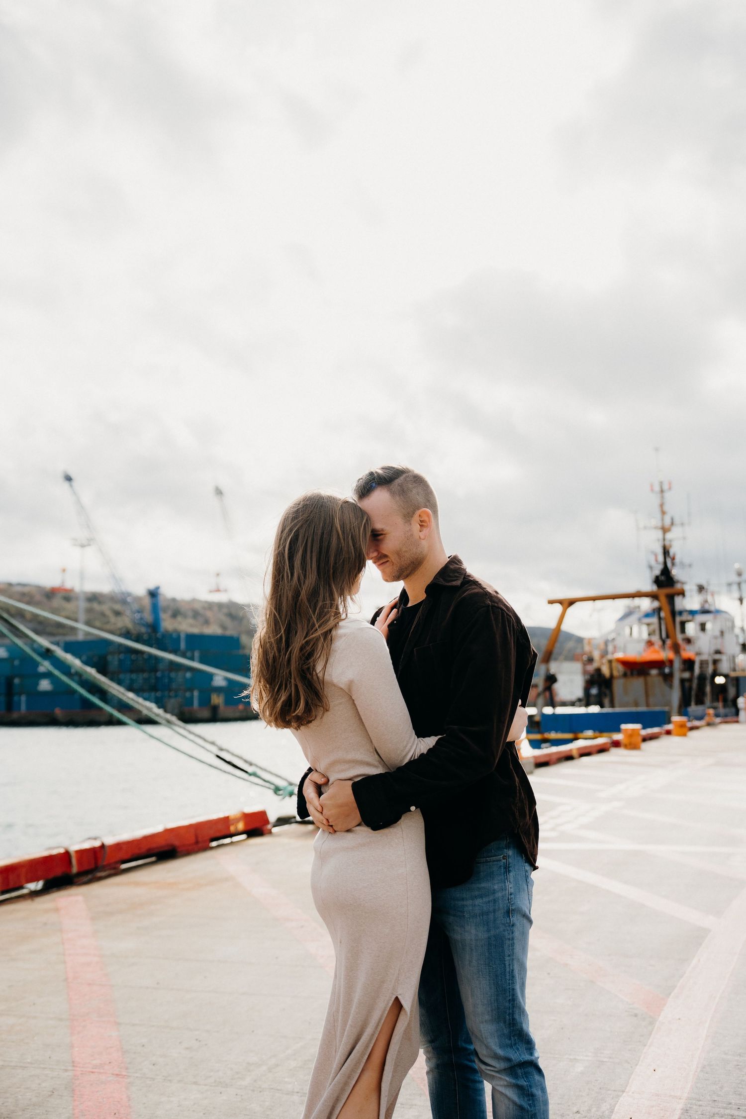 Stylish couple embracing on waterfront with boats and industrial scenery in background.