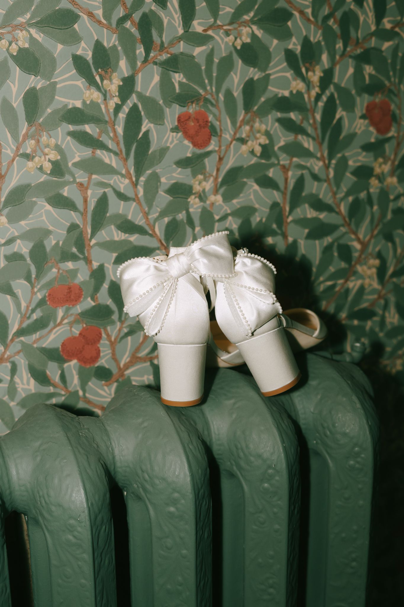 White wedding shoes with bows displayed on a vintage radiator against floral wallpaper.
