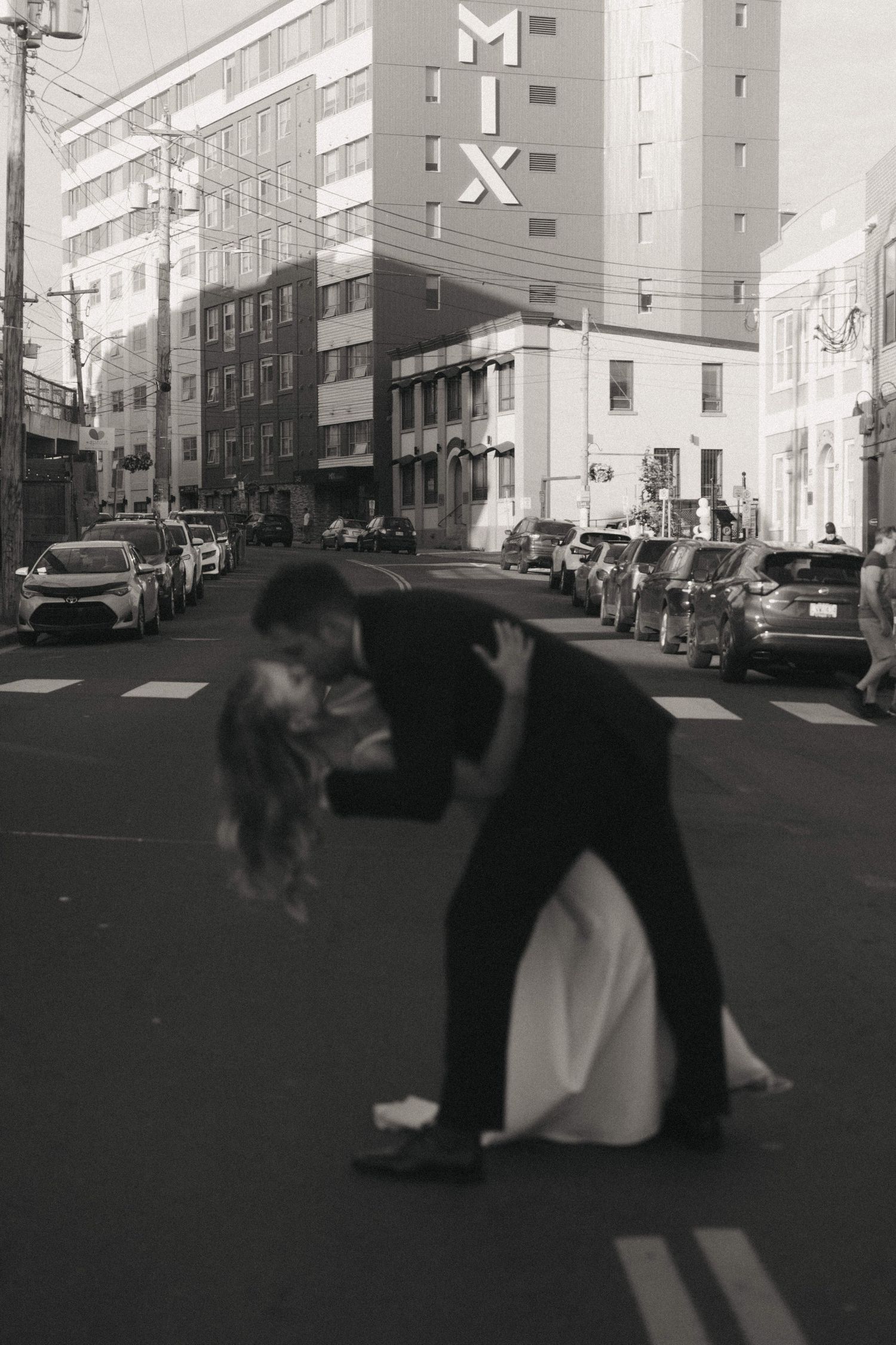 Black and white photo of dancing couple in urban setting with buildings and parked cars.