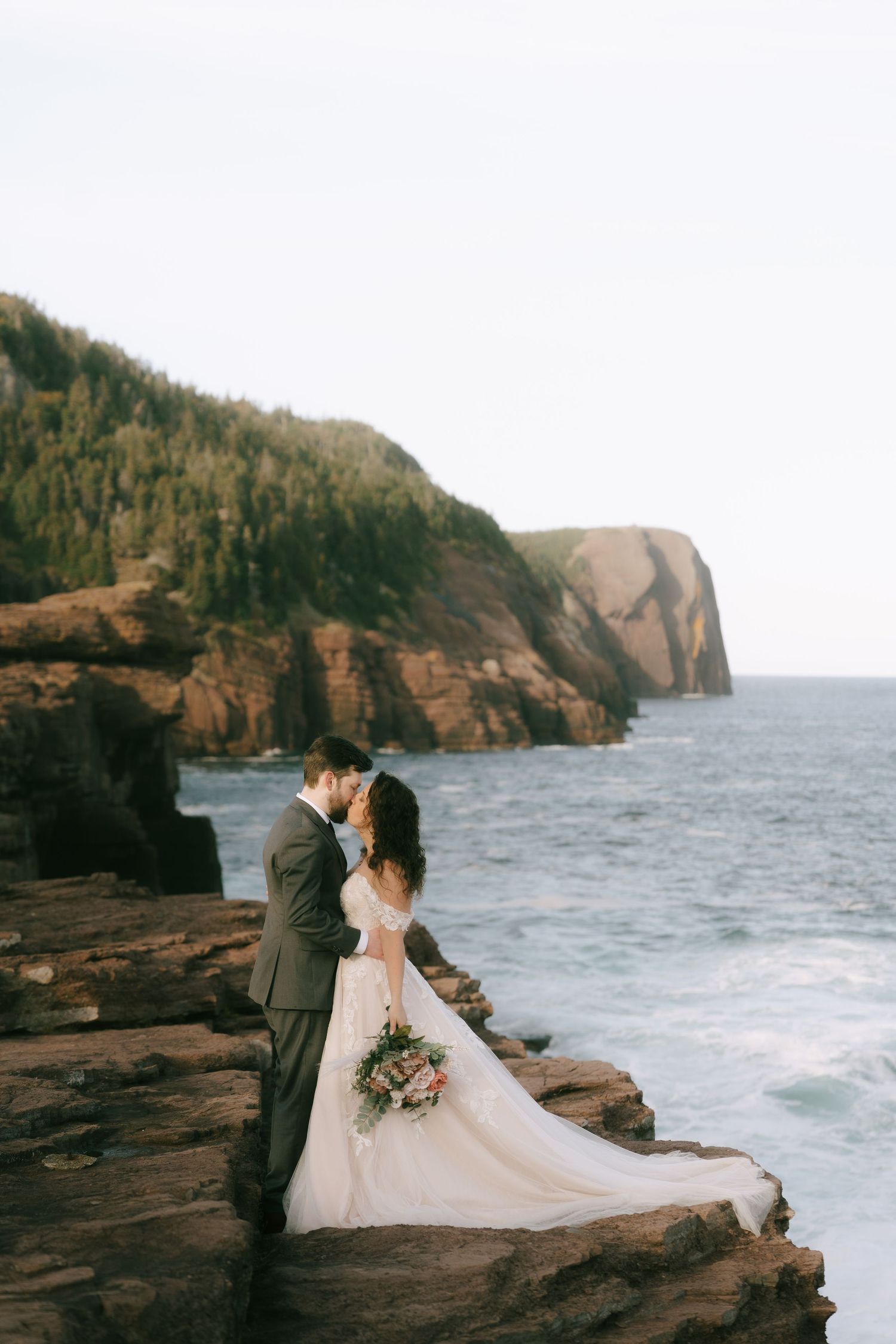 Wedding couple embracing on rocky coast with dramatic cliffs in background.