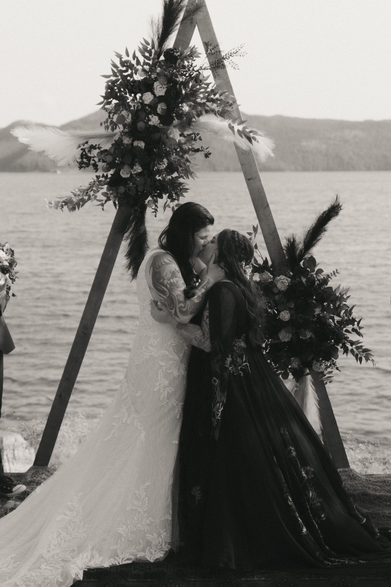 A black and white wedding ceremony photo series under an arch by the ocean with lush green florals.