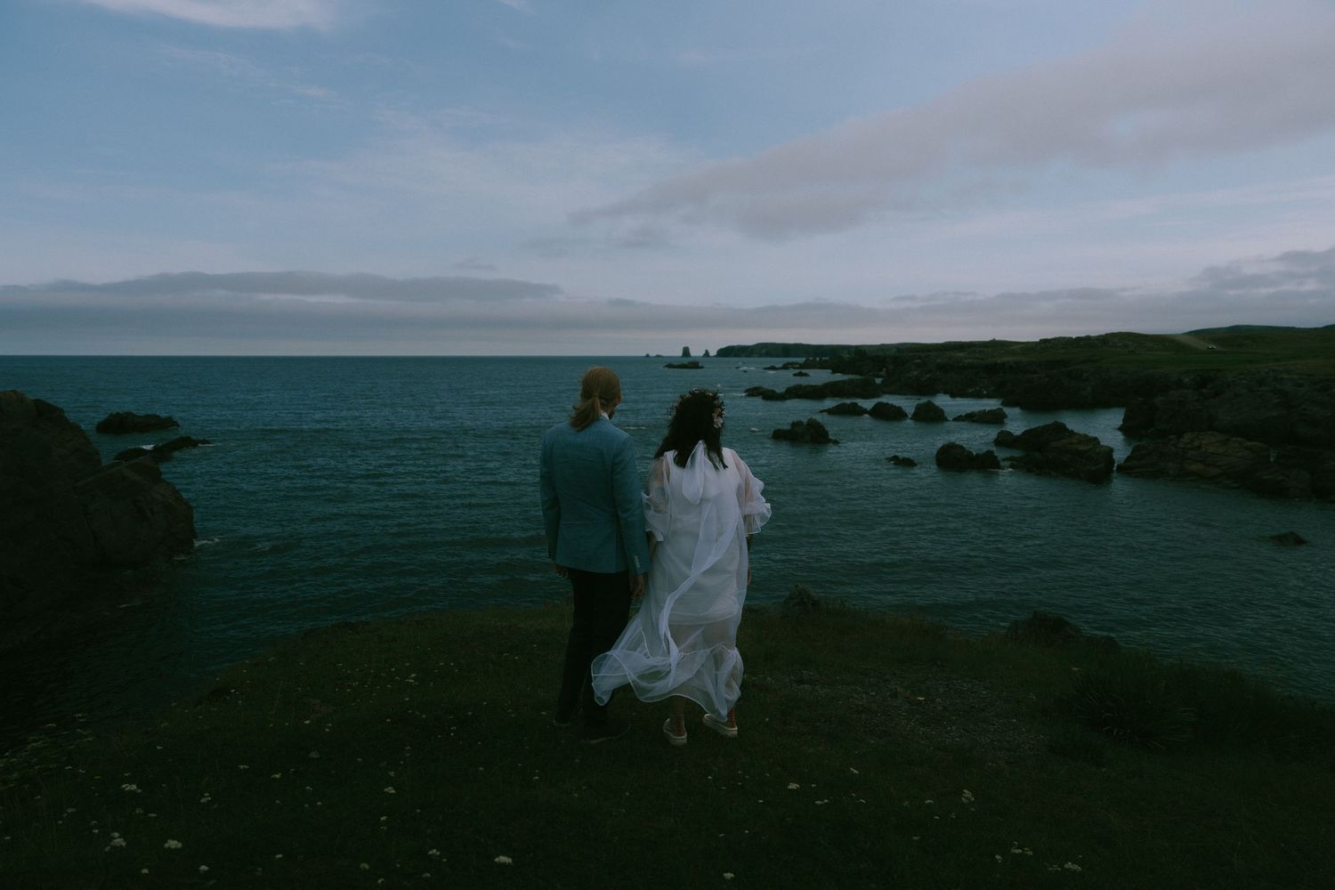 A series of dark silhouettes stand against a moody coastal landscape at dusk with dramatic cloudy skies.
