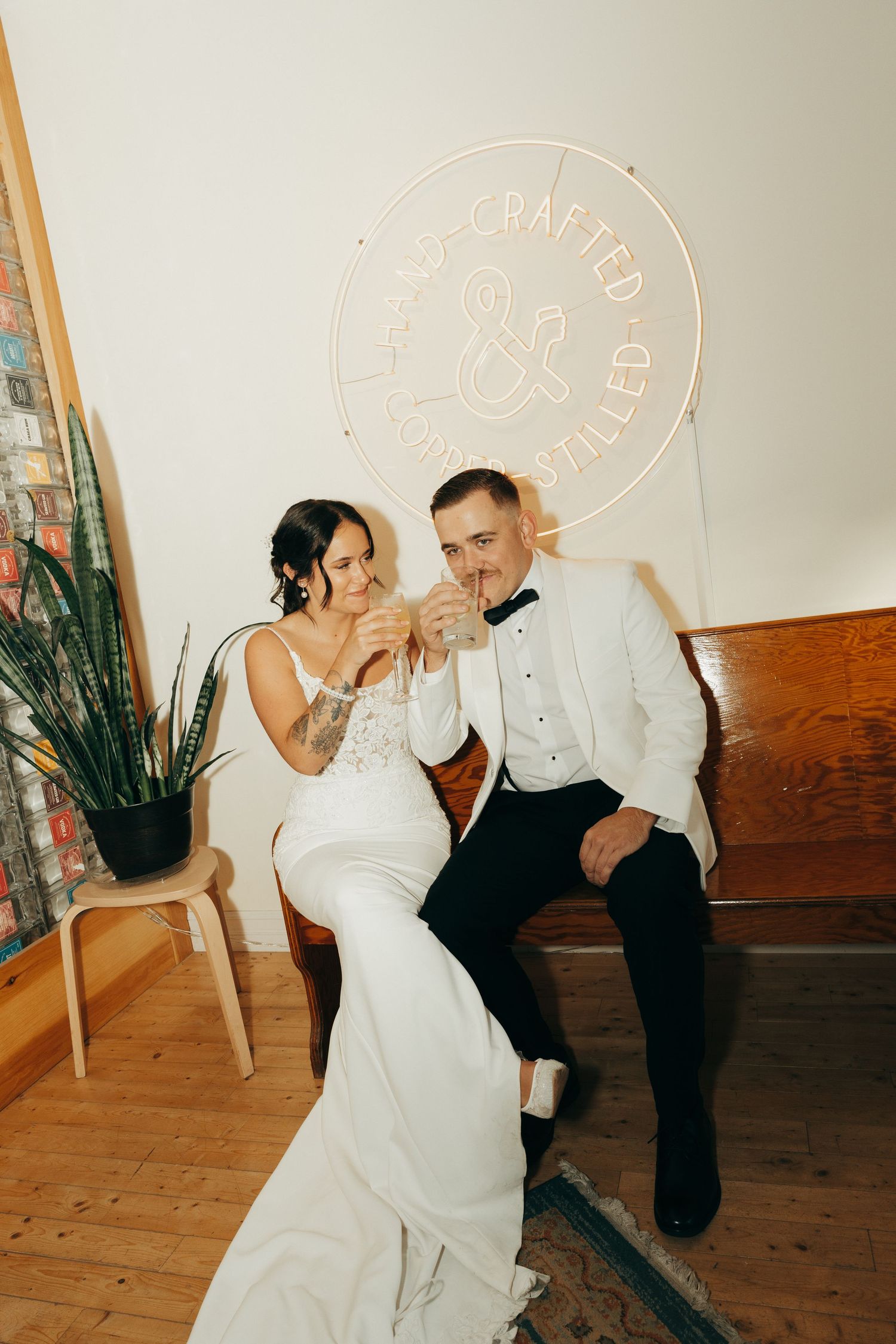 A couple in wedding attire dances playfully in a rustic room with wooden floors and neon wall signs.