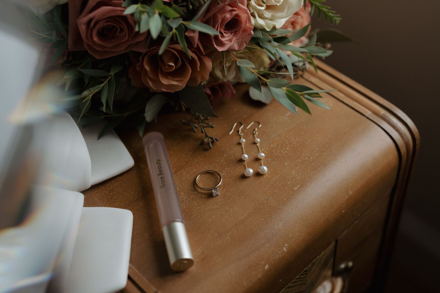 Wedding details including rose bouquet, rings, earrings and lipstick arranged on brown leather surface.