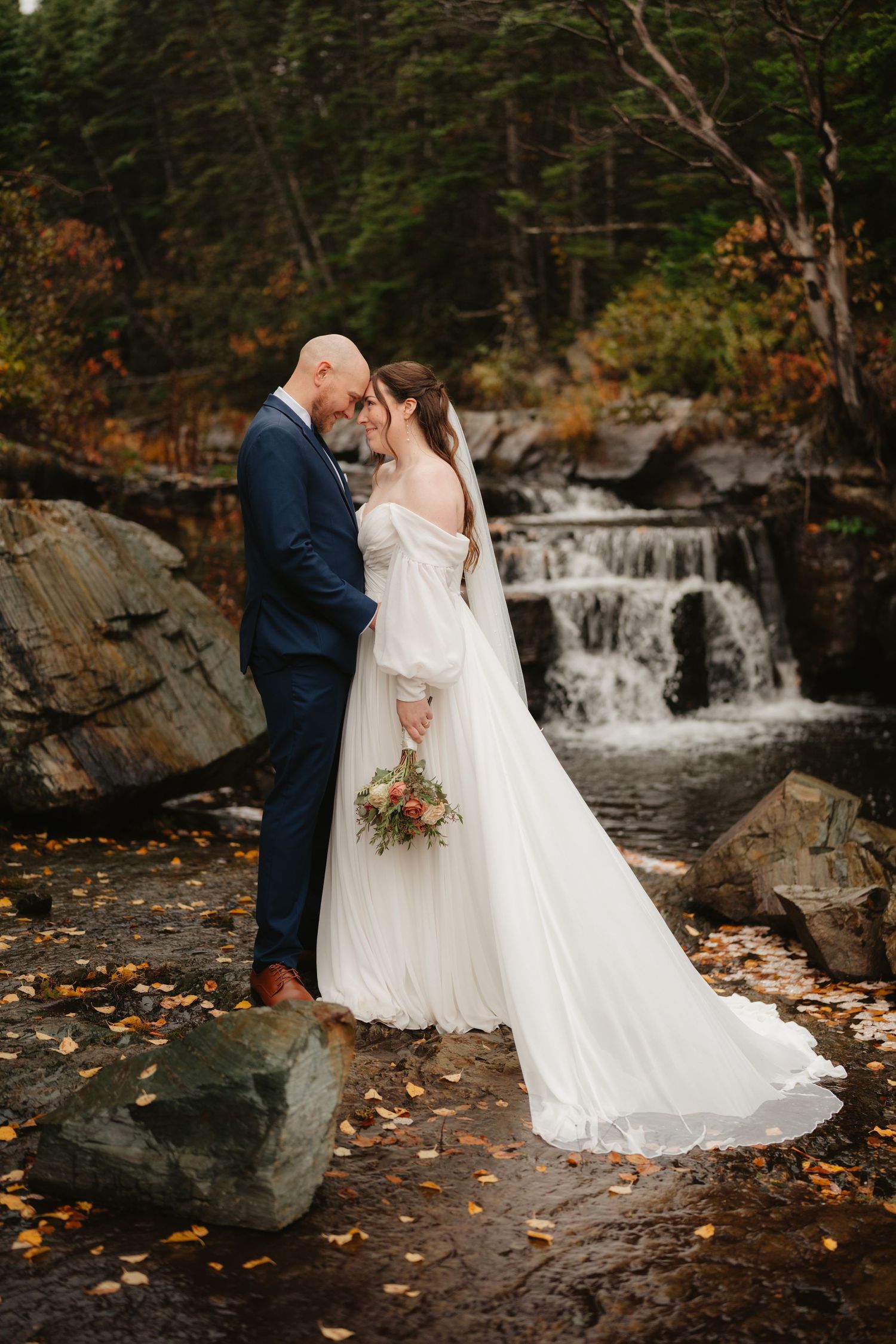 A romantic couple in wedding attire embrace near a cascading waterfall surrounded by autumn foliage and rocky terrain.