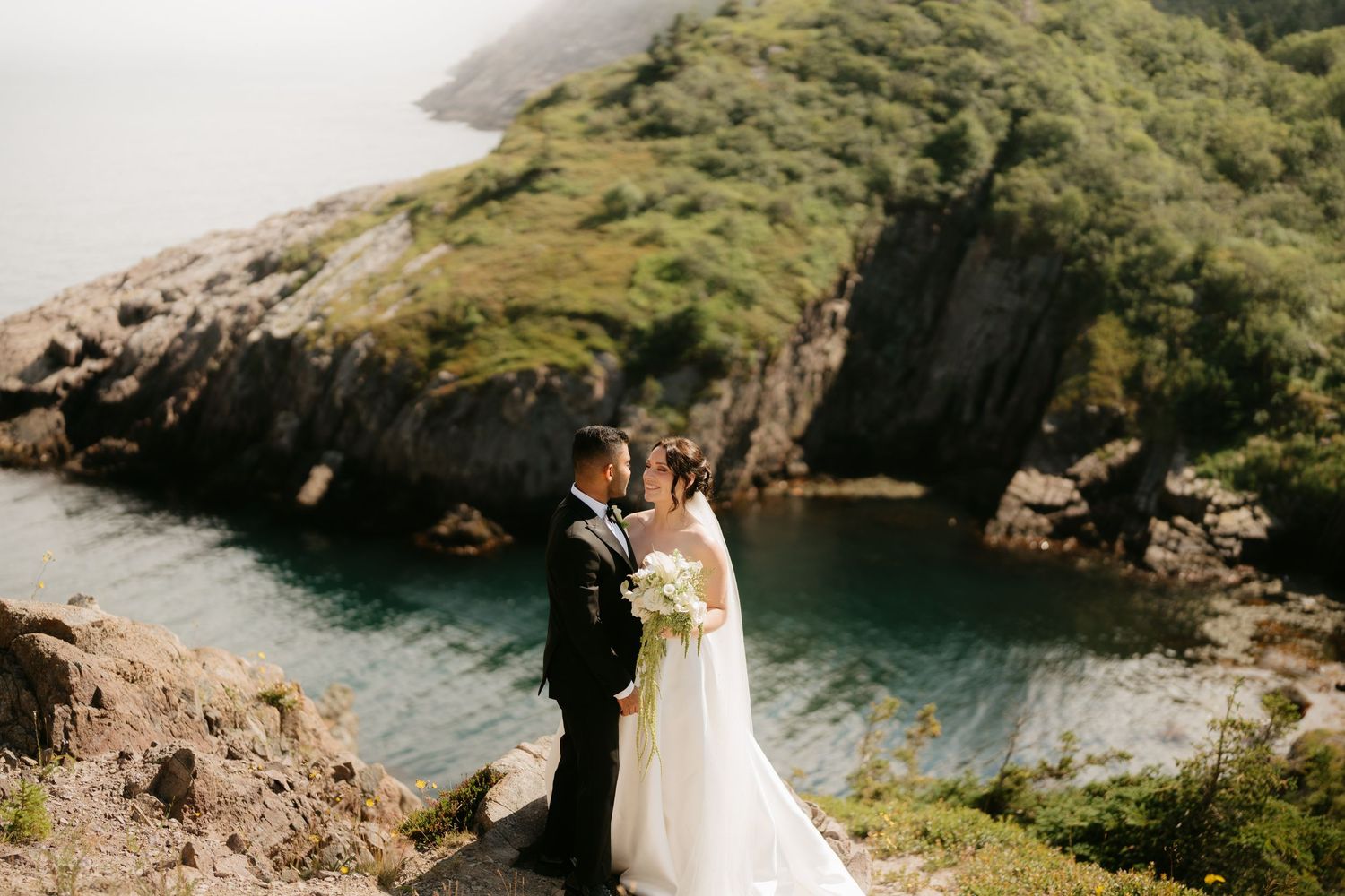 Bride and groom share romantic moment on scenic coastal cliff with turquoise water below.