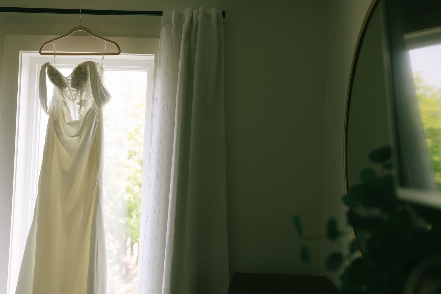 Elegant white wedding dress hanging on a hanger in front of a bright window with sheer curtains.