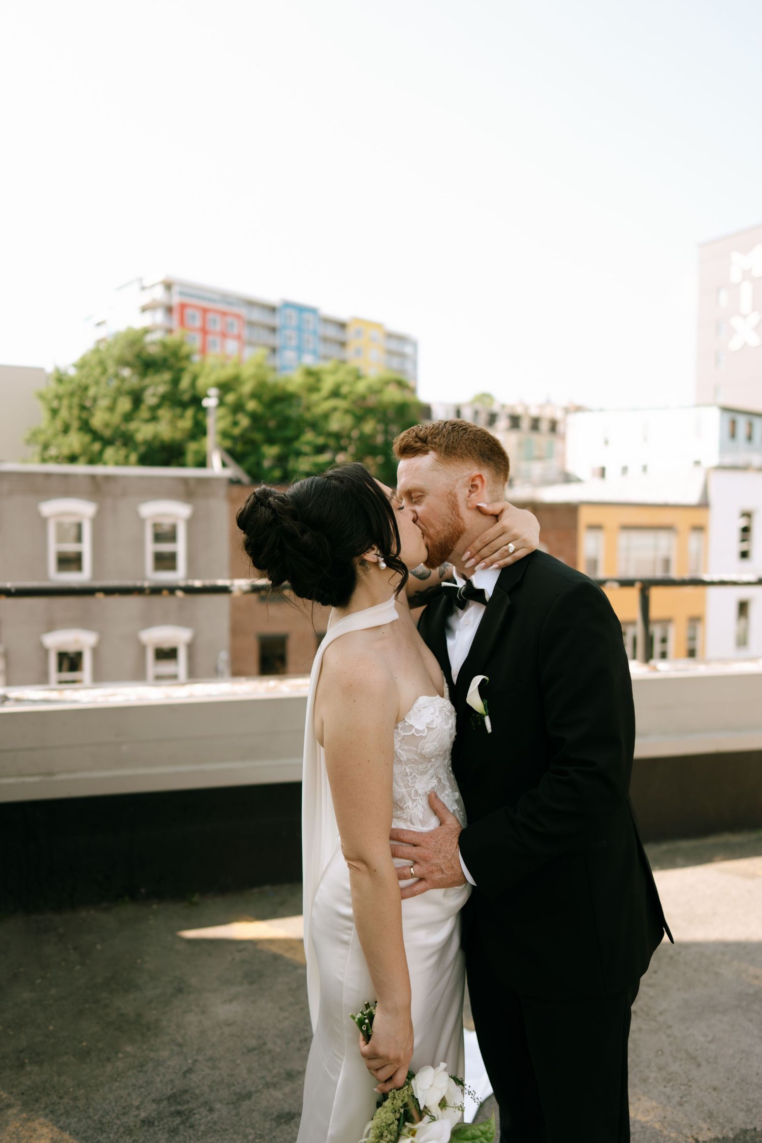 Bride and groom share a romantic rooftop kiss, she in a white halter gown, he in a black tuxedo with bow tie.