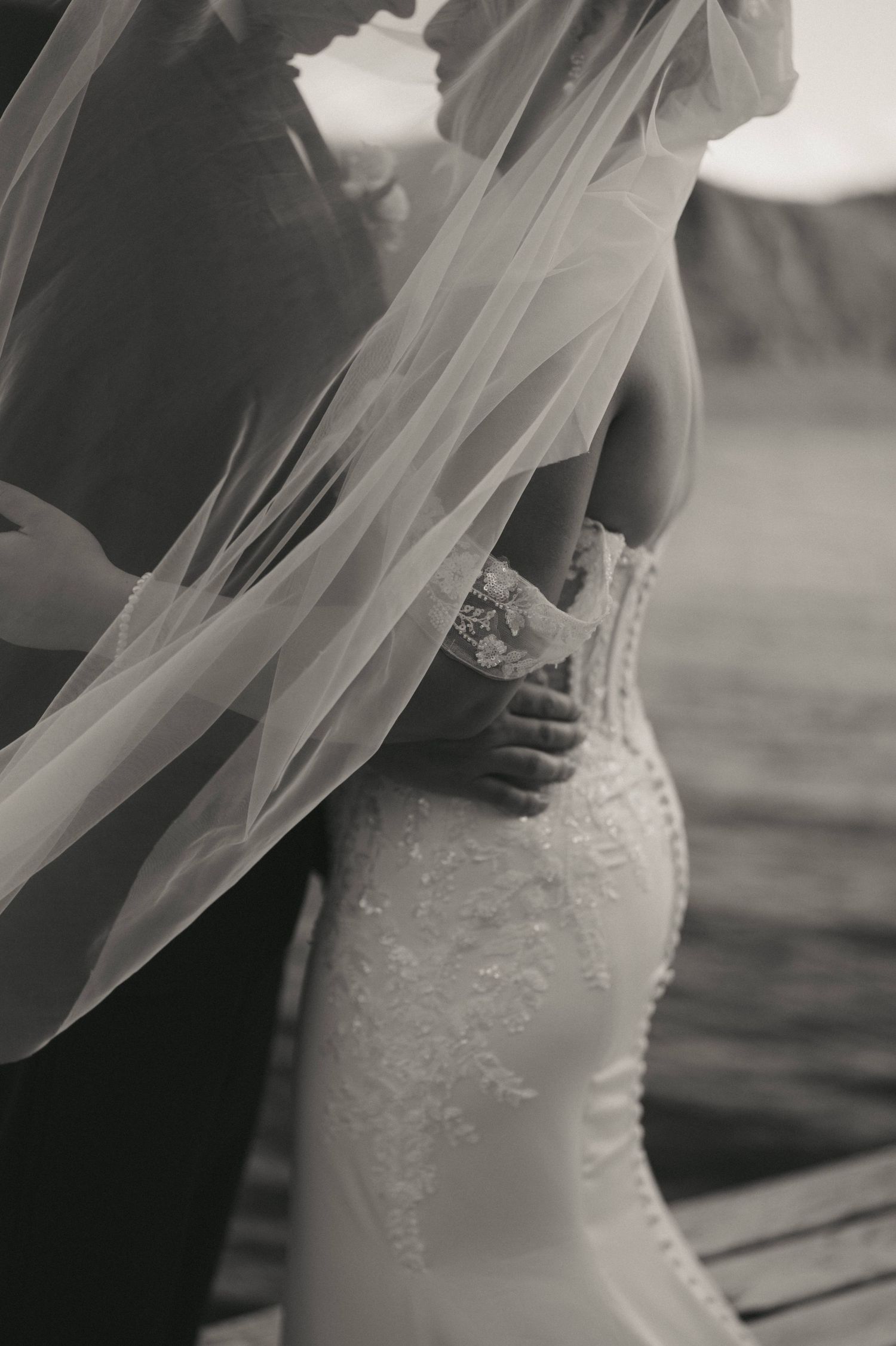 Black and white wedding photo of bride in lace gown with flowing veil, groom embracing her from behind near water.