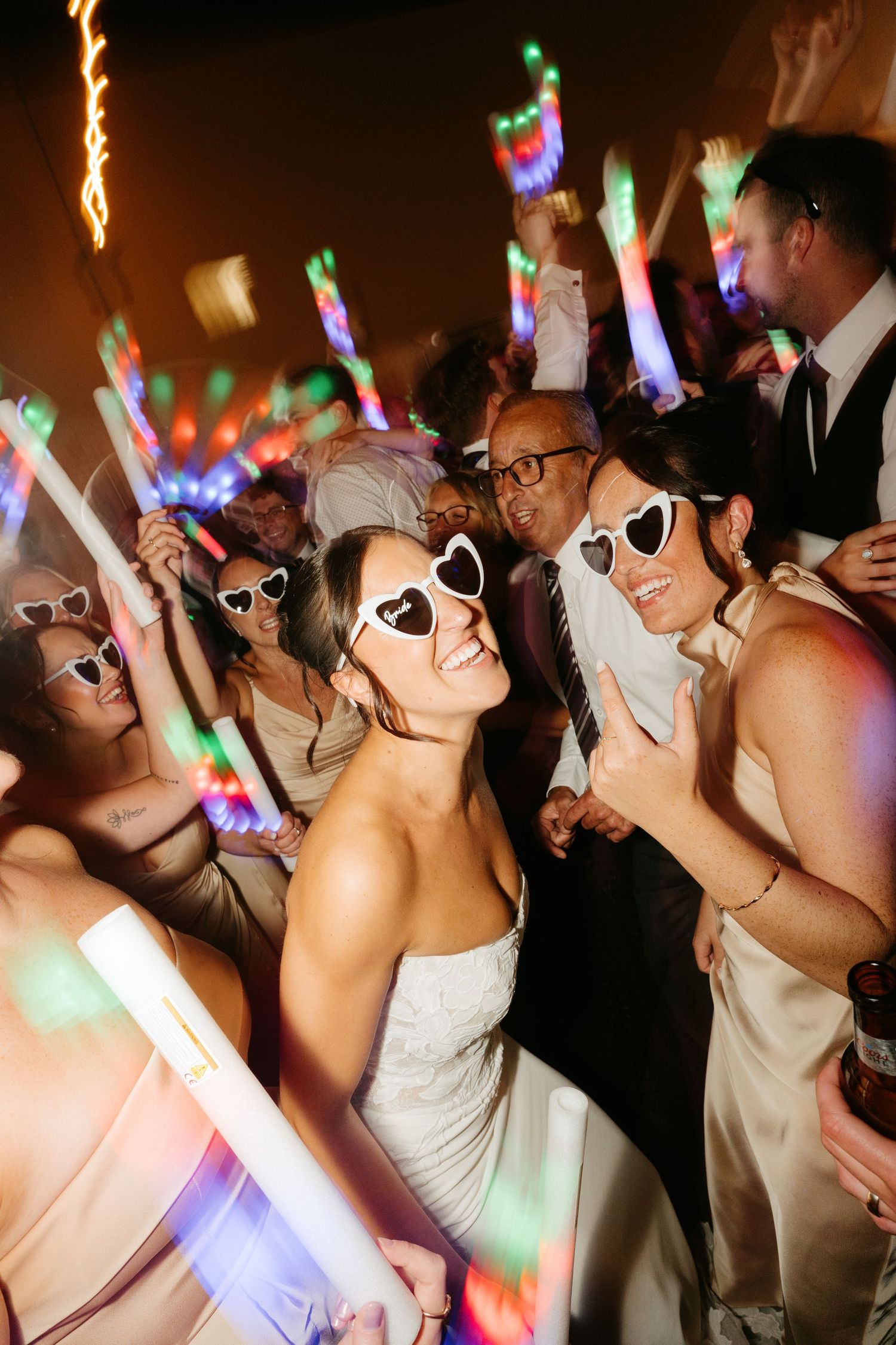 Two brides wearing heart-shaped sunglasses celebrate with glowing foam sticks at a lively wedding reception dance floor.