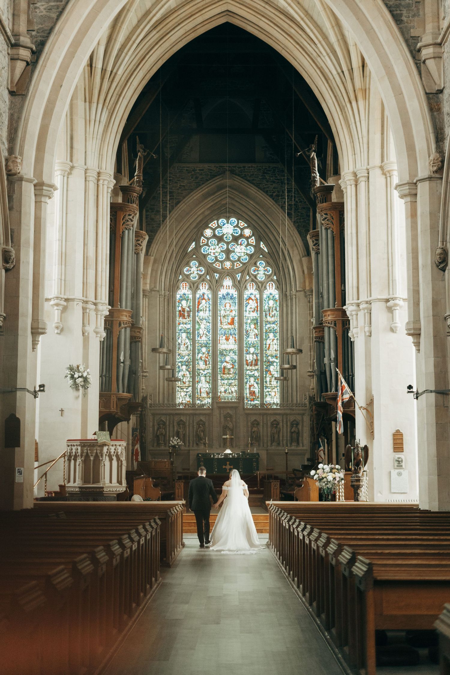 Sunlit church interior showcases majestic stone arches and stained glass windows during a wedding ceremony.
