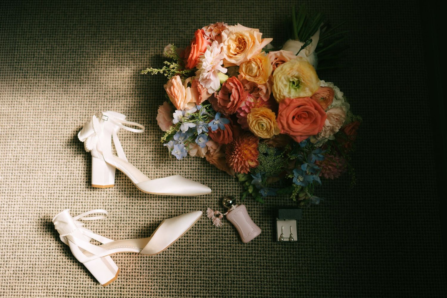 Elegant bridal shoes and coral floral arrangement displayed on neutral textured fabric.