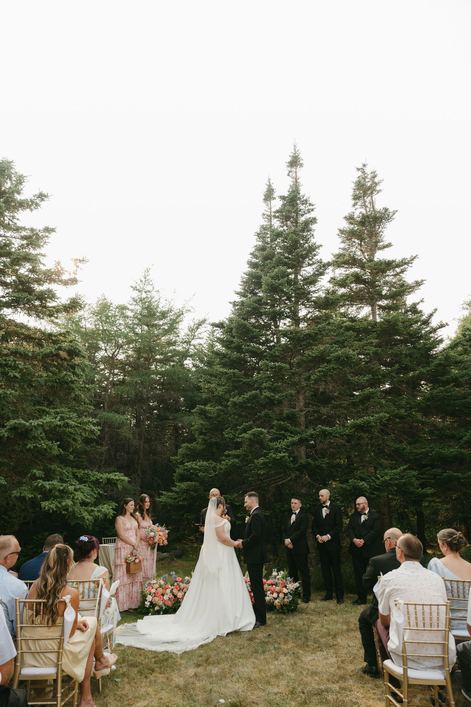 Wedding ceremony taking place in a serene outdoor setting with evergreen trees and elegant white chairs.