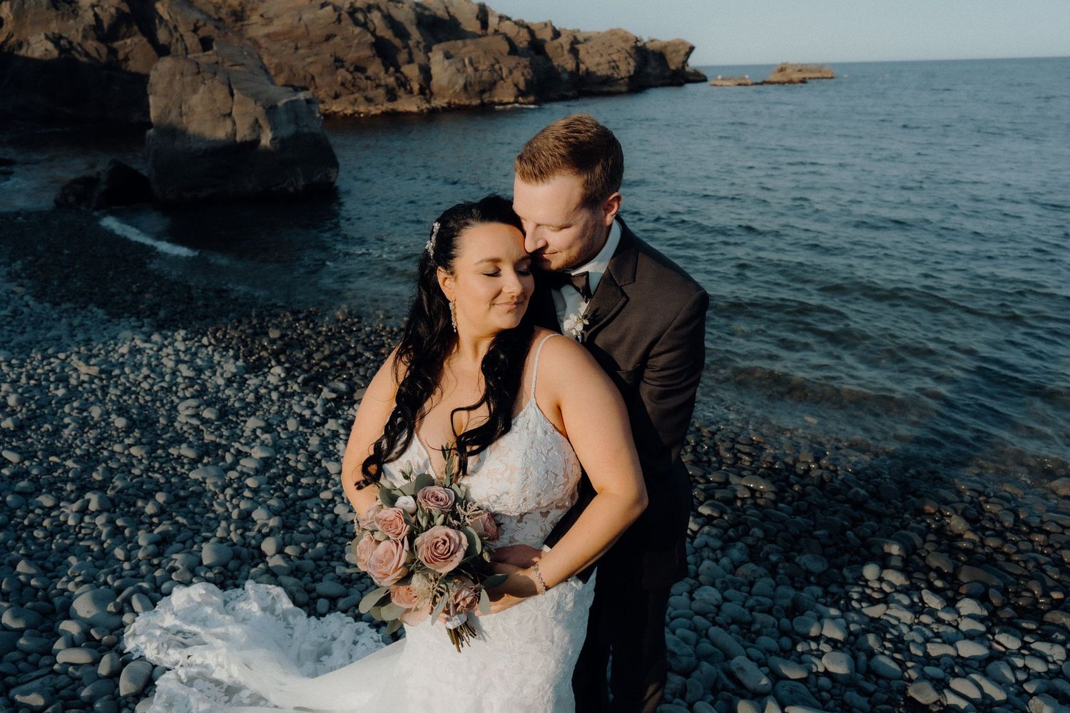 Newlyweds embrace on rocky shore with ocean and cliffs in background.