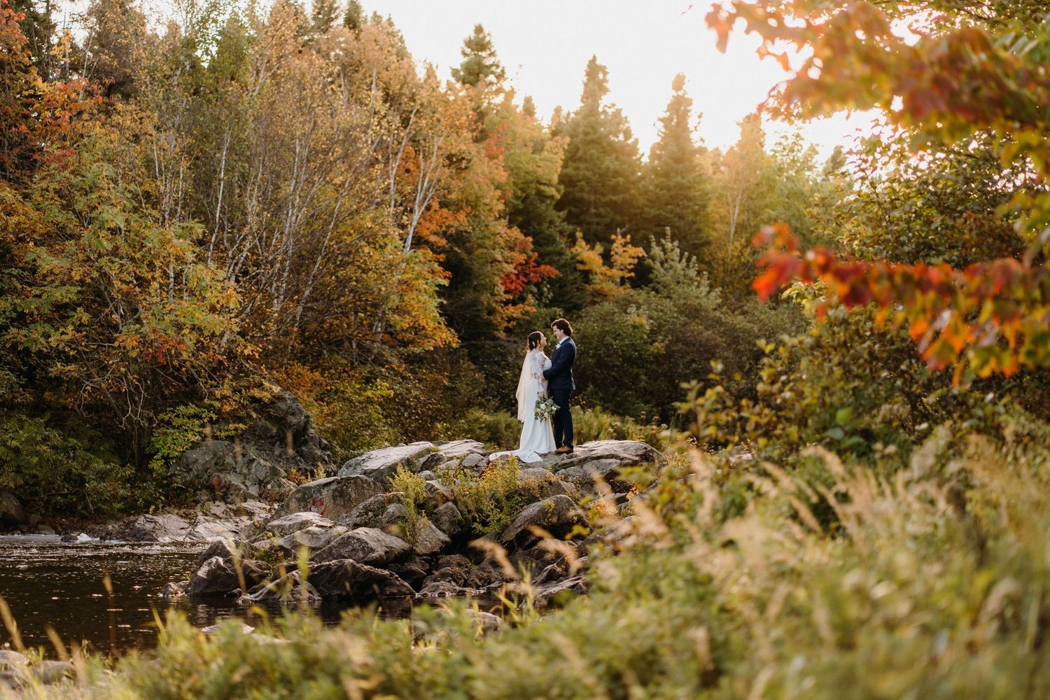 Wedding couple standing on rocky outcrop surrounded by autumn foliage.