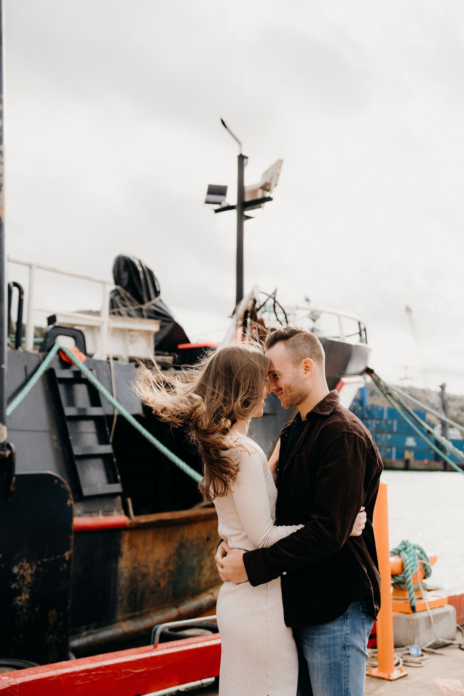 Romantic couple embraces on a dock with fishing boats in the background under cloudy skies.