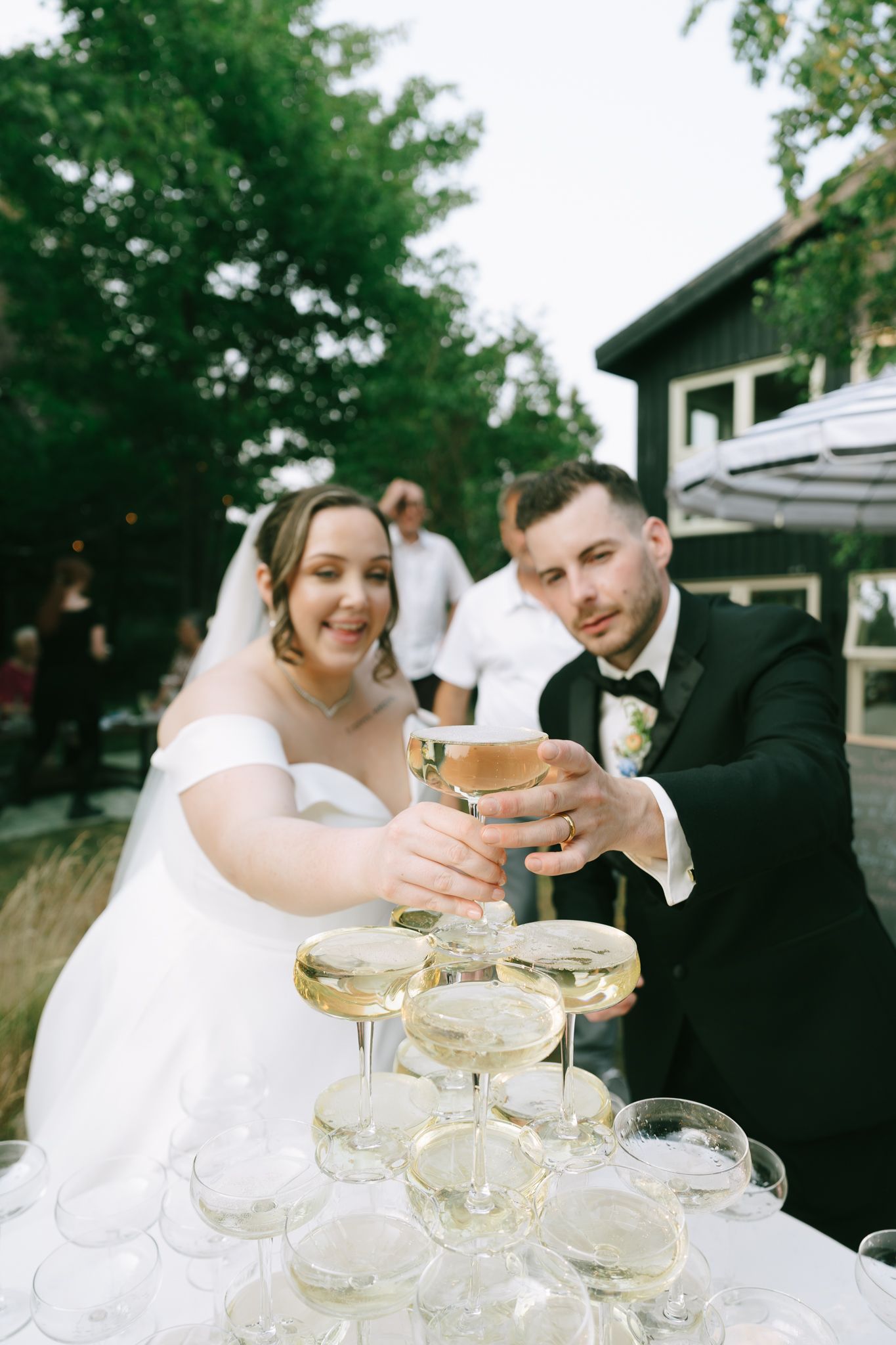 A wedding couple pouring champagne into a tower of glasses at an outdoor reception.