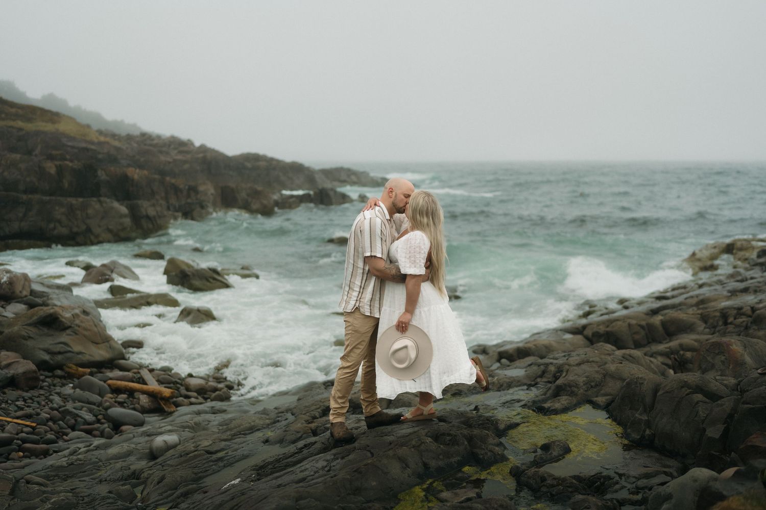 Couple in light-colored attire embraces on rocky coastal cliffs with dramatic ocean waves crashing in the background.