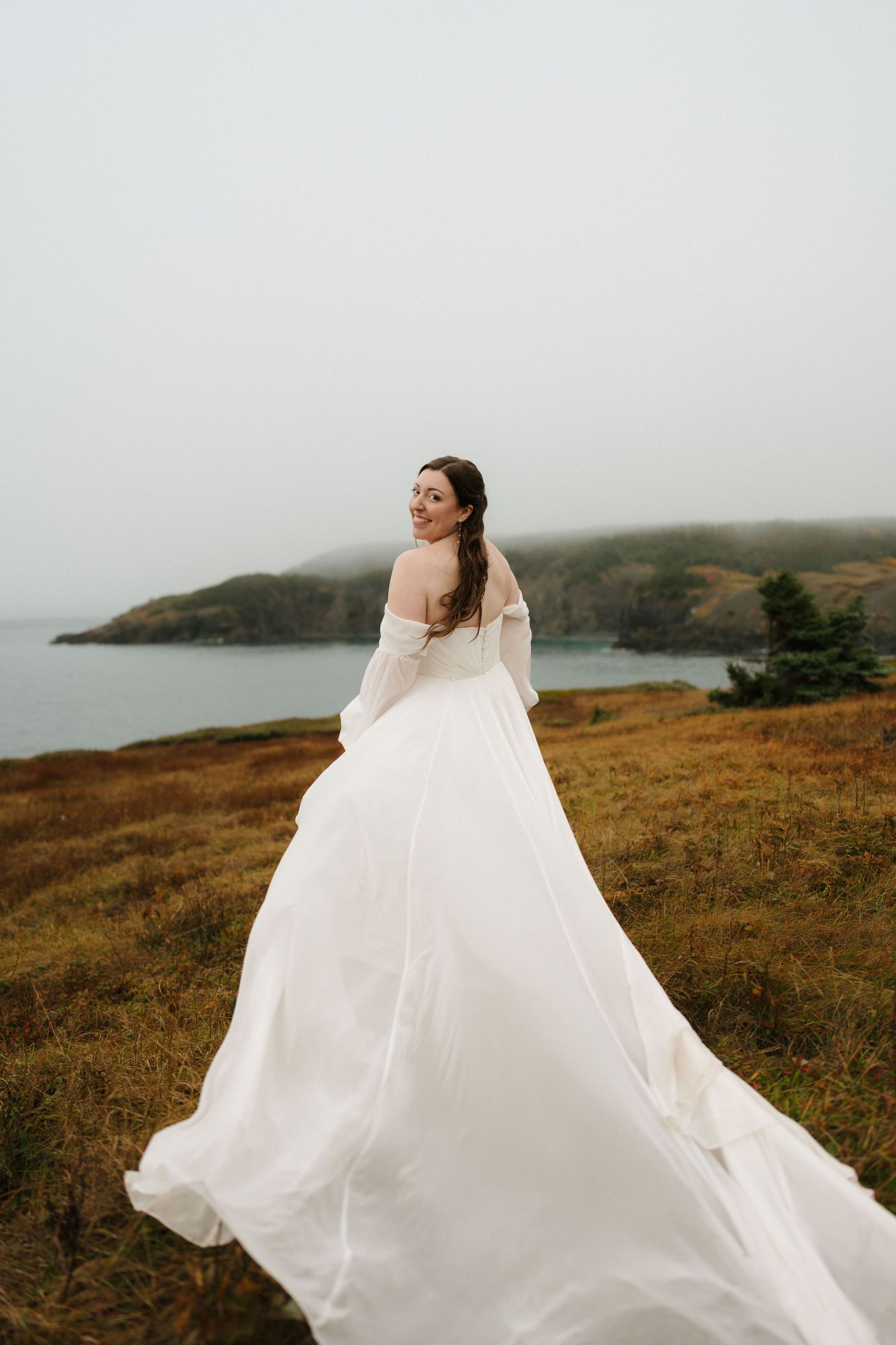 A flowing white wedding dress billows in the wind on a clifftop overlooking a misty coastal landscape.