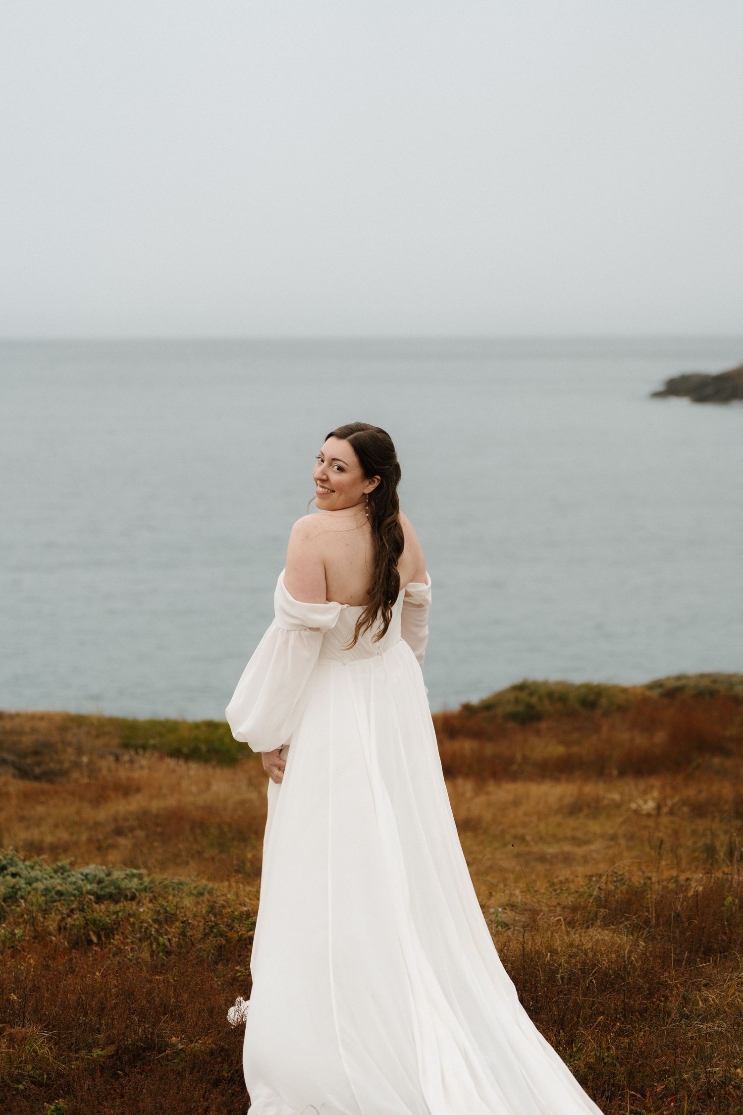 A person in a flowing white dress twirls on a coastal cliff overlooking a misty ocean view.