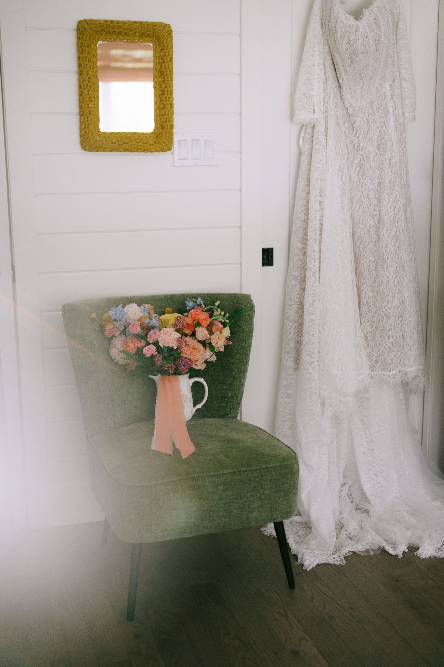 Bridal bouquet with colorful flowers in a white pitcher on a green velvet chair beside a hanging lace wedding dress.