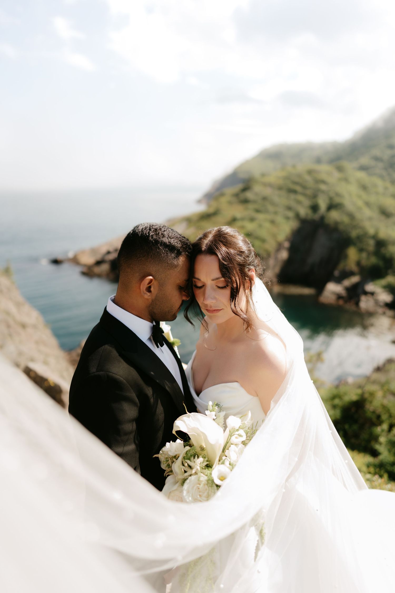 Bride and groom embrace on a cliff overlooking the ocean, bride holding white bouquet with veil flowing in the wind.