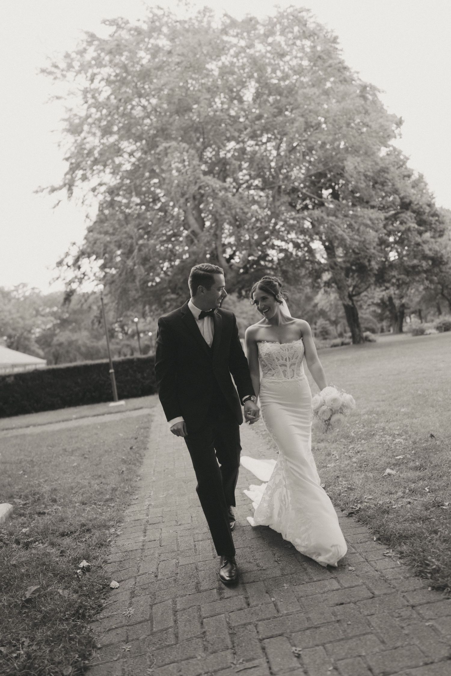 Black and white photo of a bride and groom walking hand in hand on a brick path surrounded by lush trees.