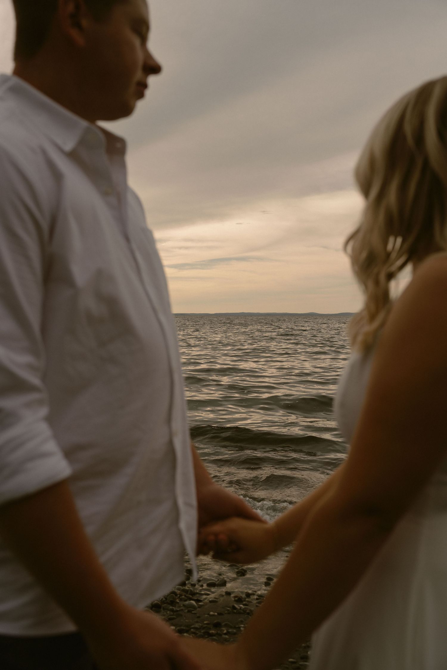 A couple stands together at sunset by the ocean, with dramatic cloudy skies and waves in the background.