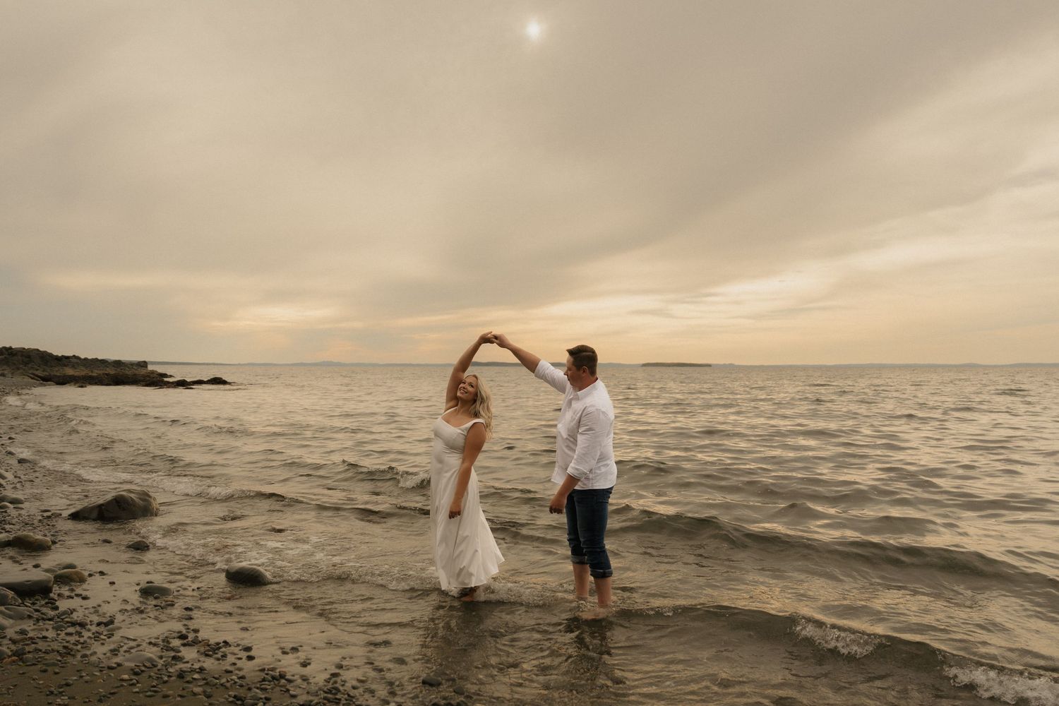 A couple walks along a scenic beach at sunset as waves crash against the shore while holding hands.