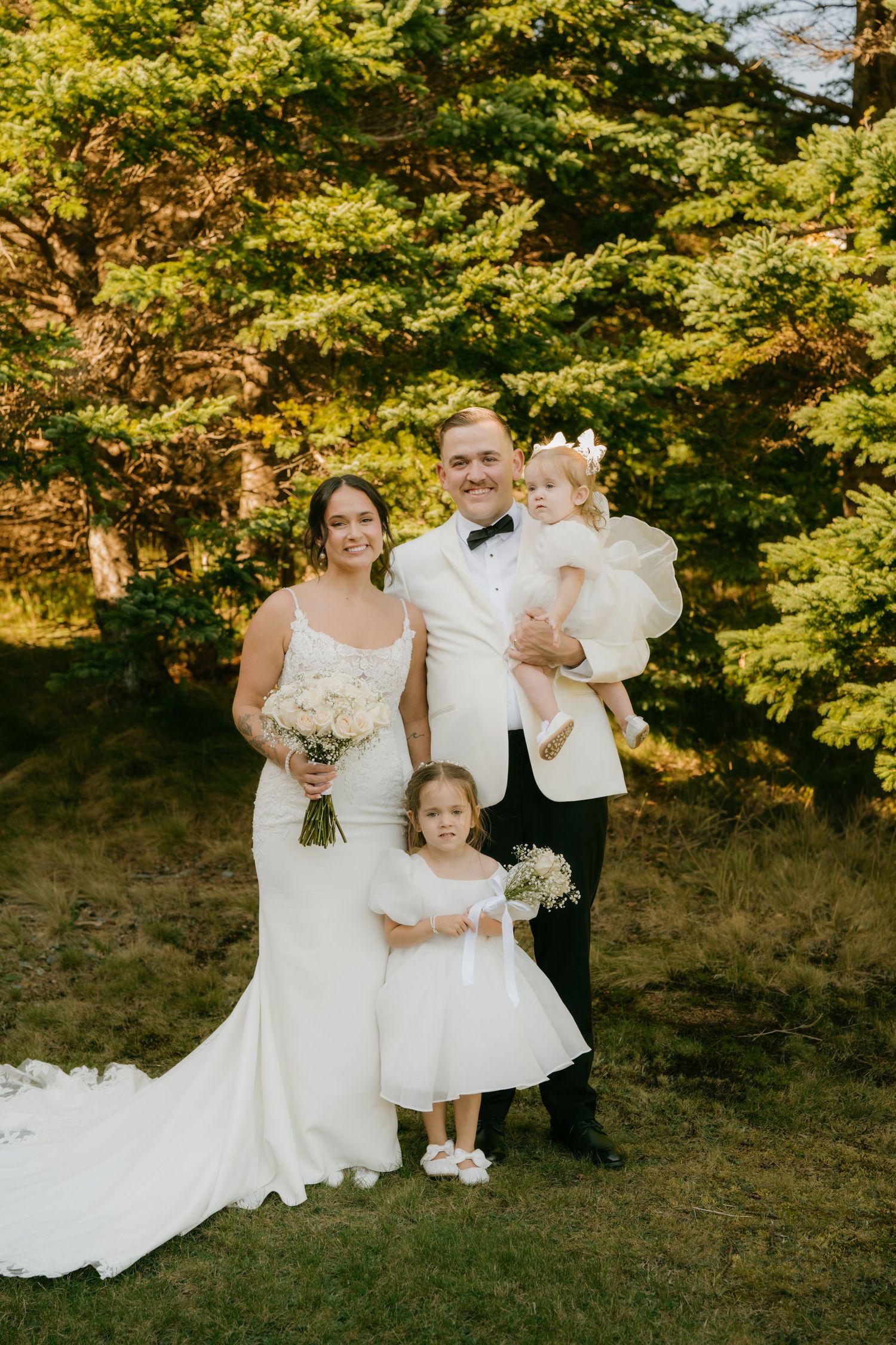 A wedding party poses outdoors in formal white attire against a backdrop of lush green trees on a sunny day.