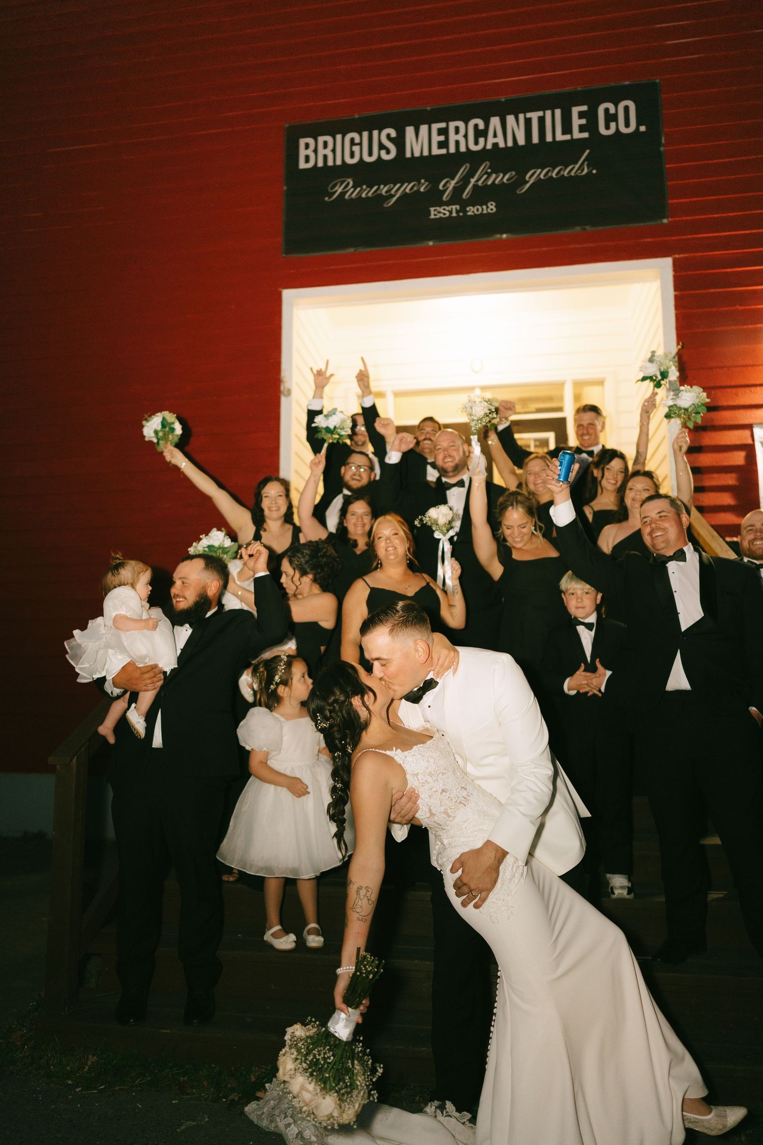 Wedding guests dance and celebrate at a rustic reception venue with a vintage Briggs Mercantile Co. sign visible.
