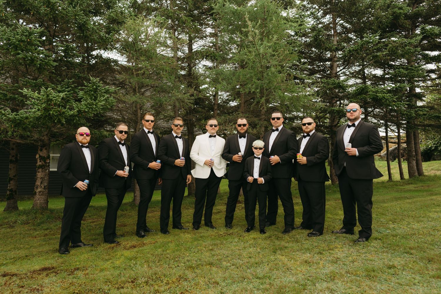 A wedding party in formal black and white attire poses in a line formation on a grassy lawn with trees in the background.