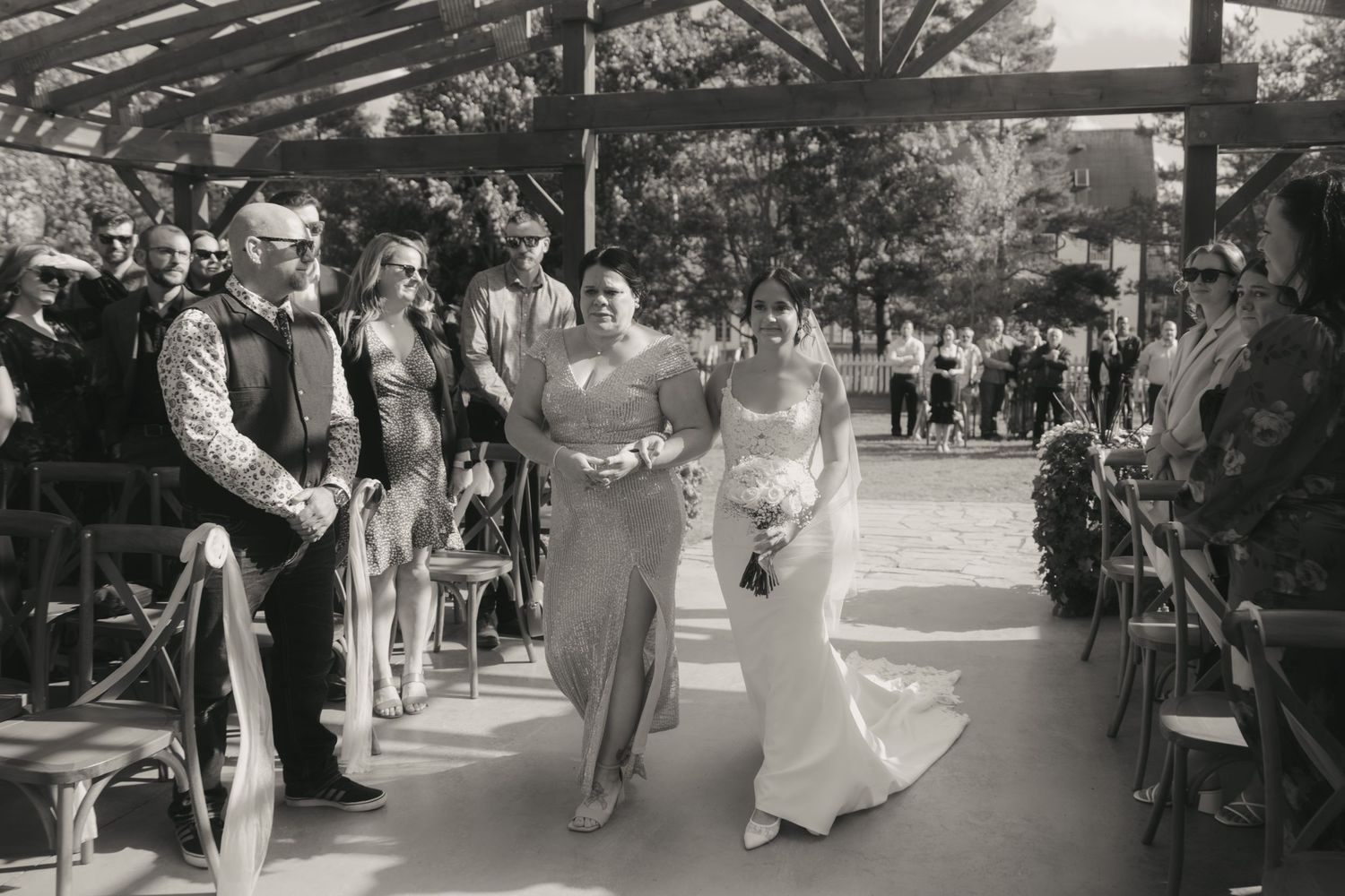 Black and white wedding ceremony photo showing a couple walking down an outdoor aisle lined with guests under a pergola.