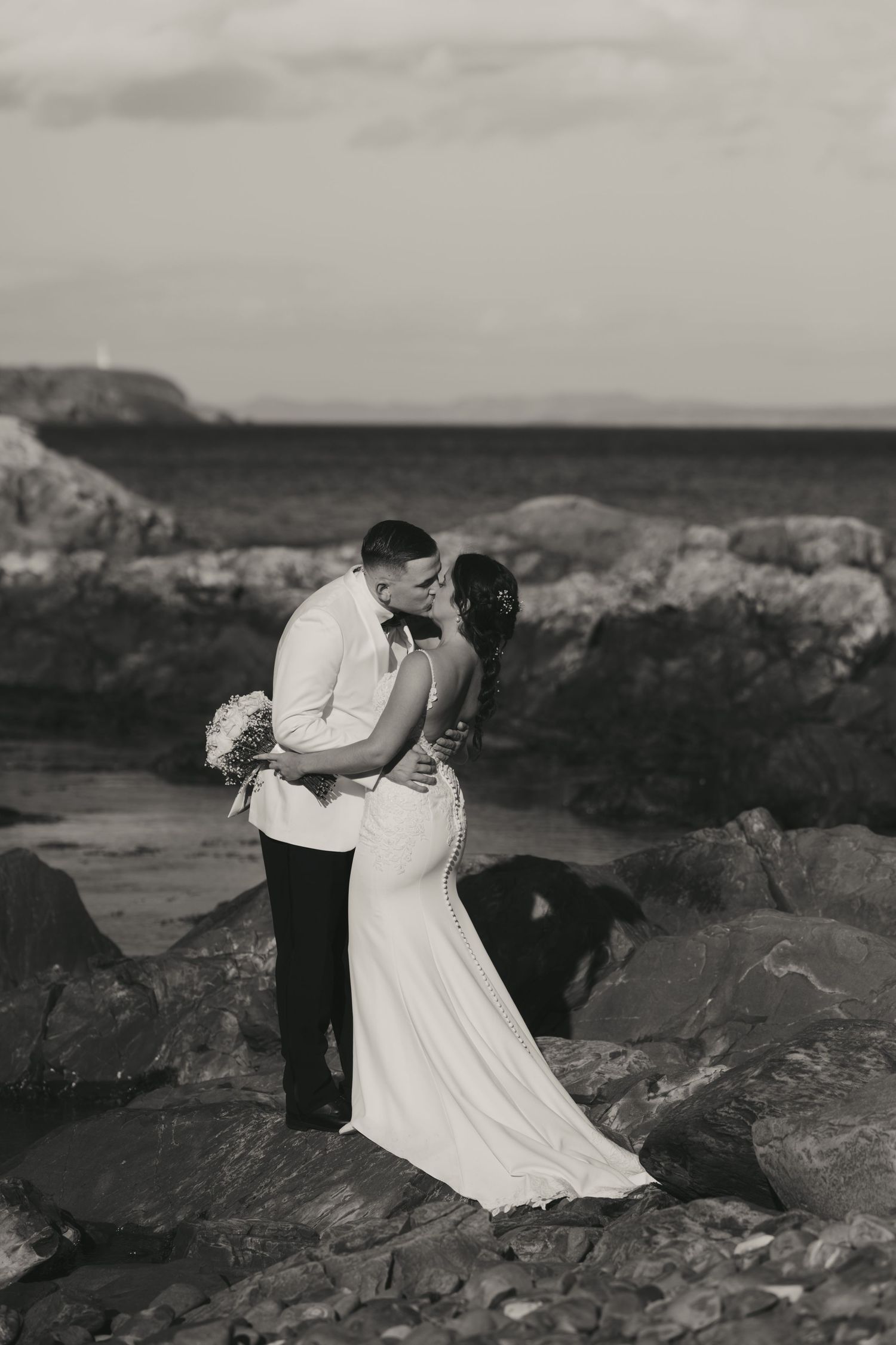 A romantic black and white wedding photo series showing a couple embracing on rocky coastal cliffs at sunset.