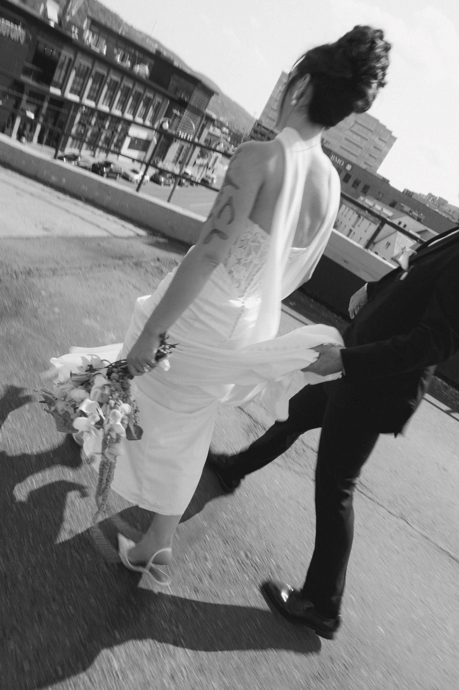 Black and white photo of a bride in white dress holding bouquet walking with groom on urban street.