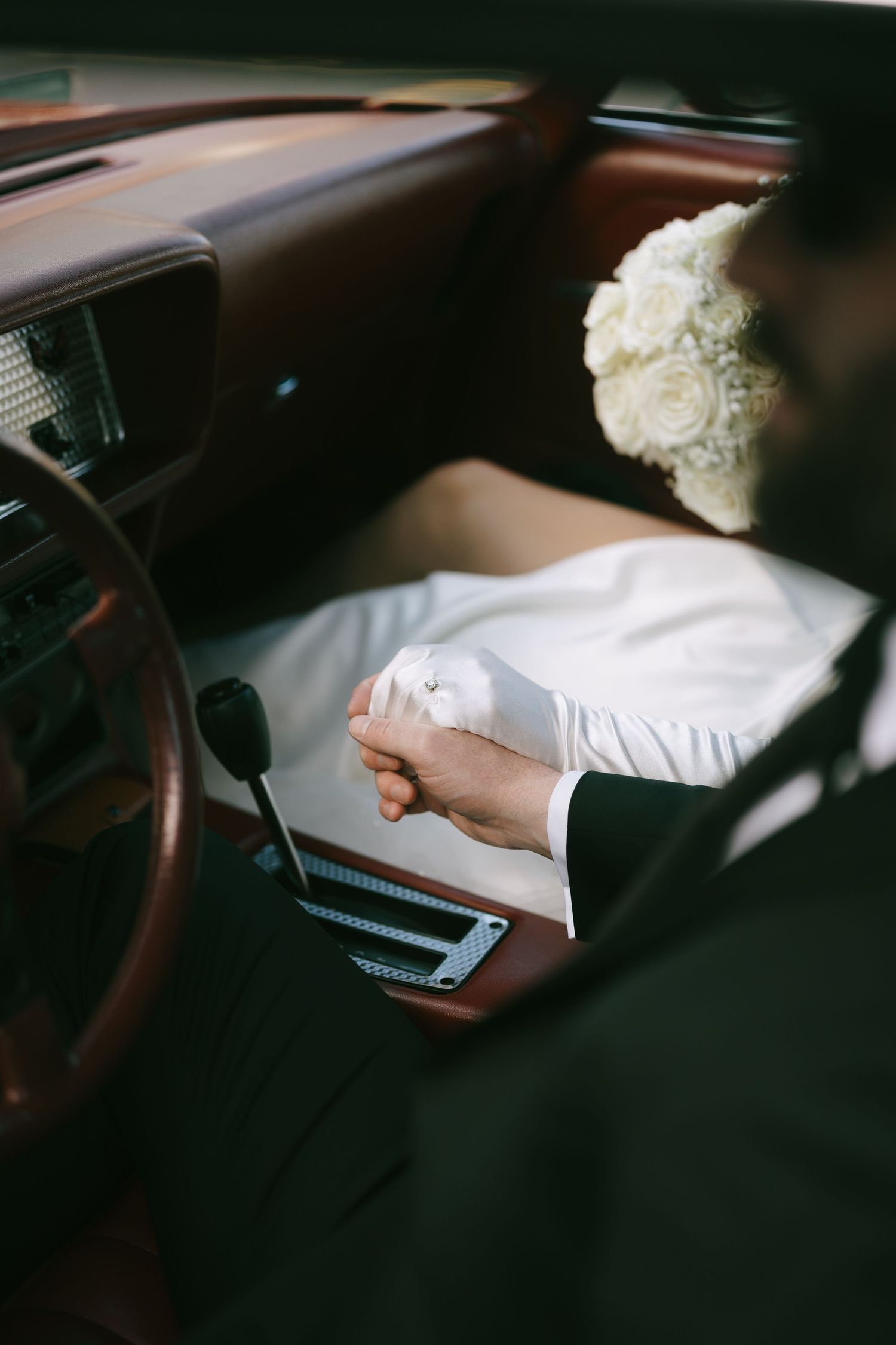 Bride and groom holding hands in a vintage car, with a white rose bouquet visible on the dashboard.