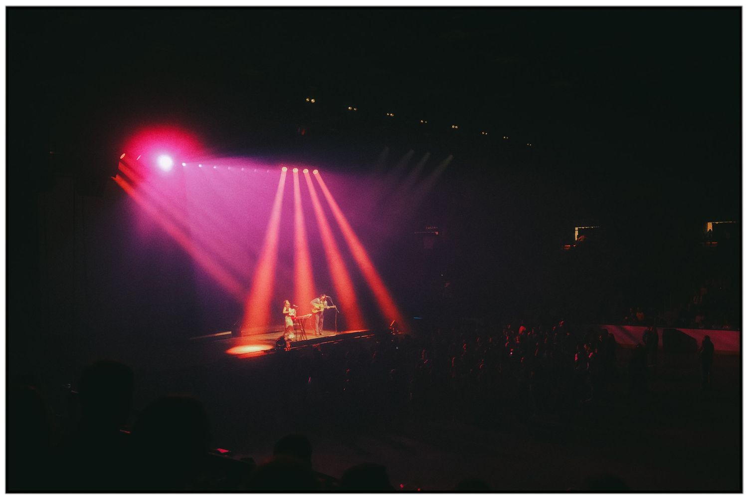 Pink and red stage lights create dramatic beams through fog during a concert performance.