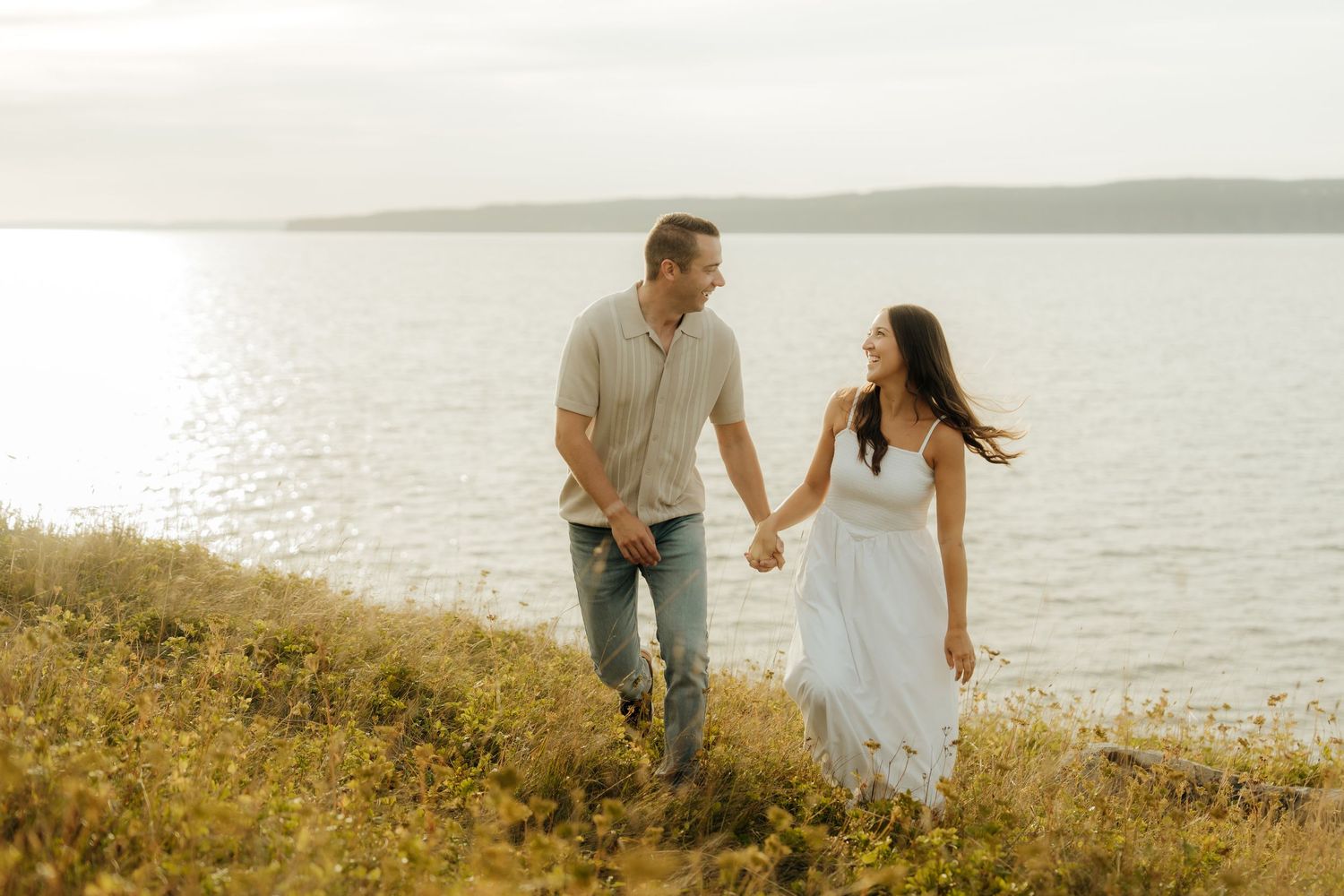 A couple walks hand in hand along a grassy coastal cliff at sunset in a romantic summer setting.