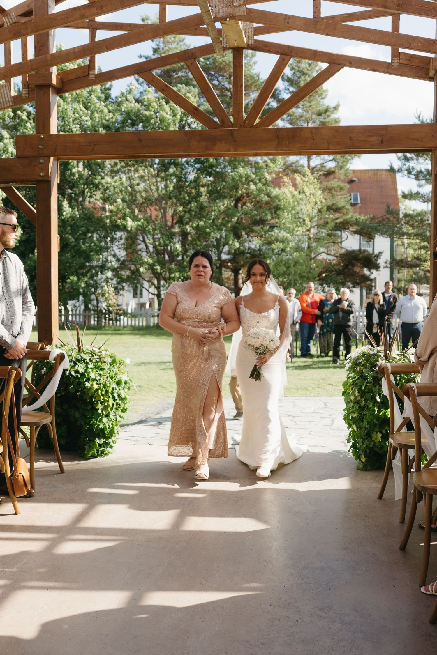 A wedding party enters a rustic wooden pavilion decorated with white drapes and greenery during an outdoor ceremony.
