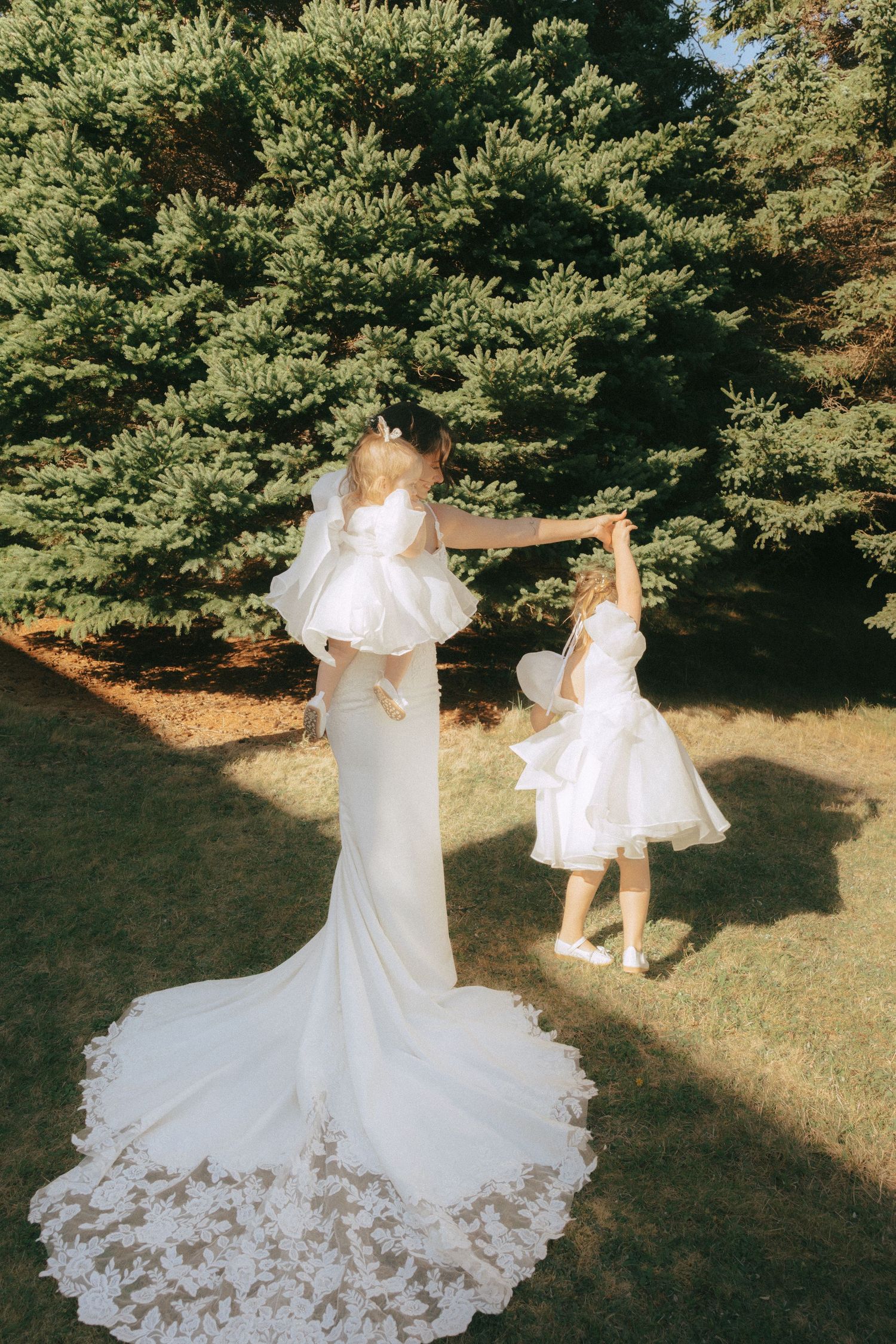 Two figures in flowing white dresses dance together on a grassy lawn surrounded by evergreen trees.
