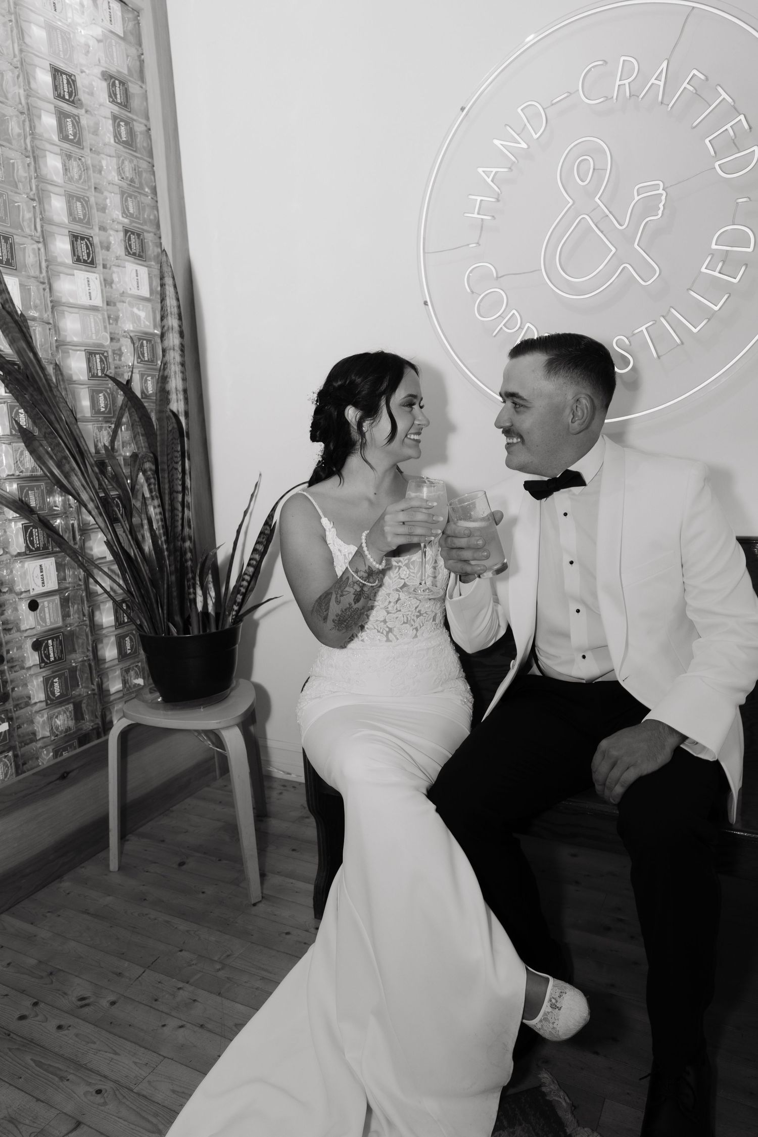 A couple in wedding attire shares a drink together while sitting in front of a circular logo on a white wall.