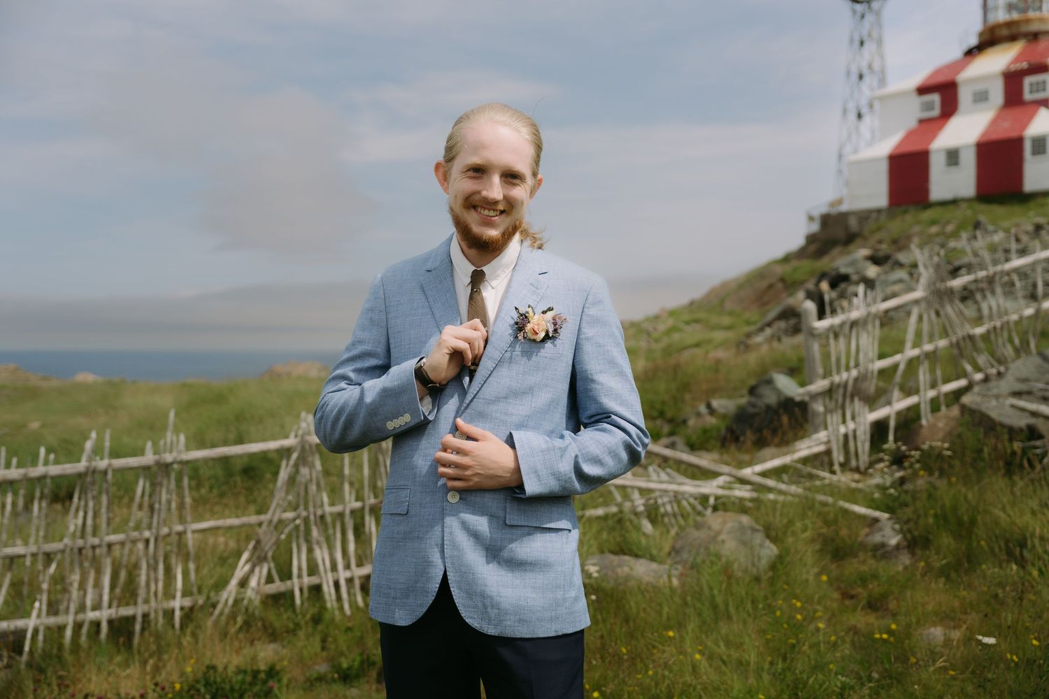 A person in a light blue suit jacket stands near wooden fences on a grassy coastal hillside.